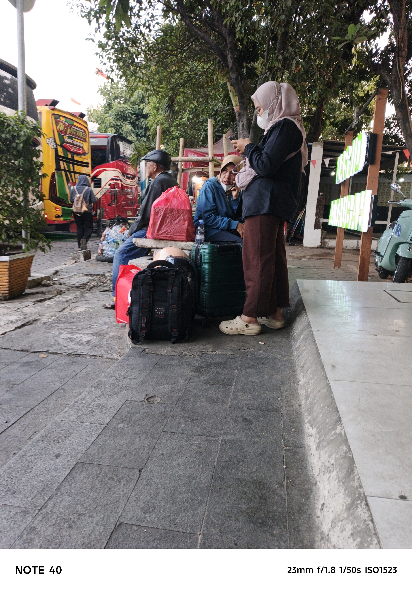 A medium-full shot captures a street scene in North Jakarta, Indonesia, focusing on a group of people and luggage next to a bus stop. The foreground features a gray concrete sidewalk with cracked tiles and debris. To the right, a white concrete curb marks the edge of a raised area. Behind the curb, a mint-green scooter is parked. Further back, two lit signs with green lettering are mounted on wooden posts.
In the midground, three individuals are seated on a low bench. The person on the left, wearing a black cap, dark jacket, and blue jeans, has a red plastic bag on their lap. The person in the middle, wearing a blue jacket and tan head covering, is looking away. The person on the right, wearing a light pink hijab, a dark jacket, and brown pants, is holding a smartphone. Several pieces of luggage are present: a black backpack is in the lower center, a green suitcase is to its right, and a large red plastic bag is behind the person on the left. A partially visible red and white bus is parked behind the seated individuals, with a yellow and red bus to its left. Trees with green foliage line the background. The scene appears to be a waiting area, likely at a bus terminal or station.