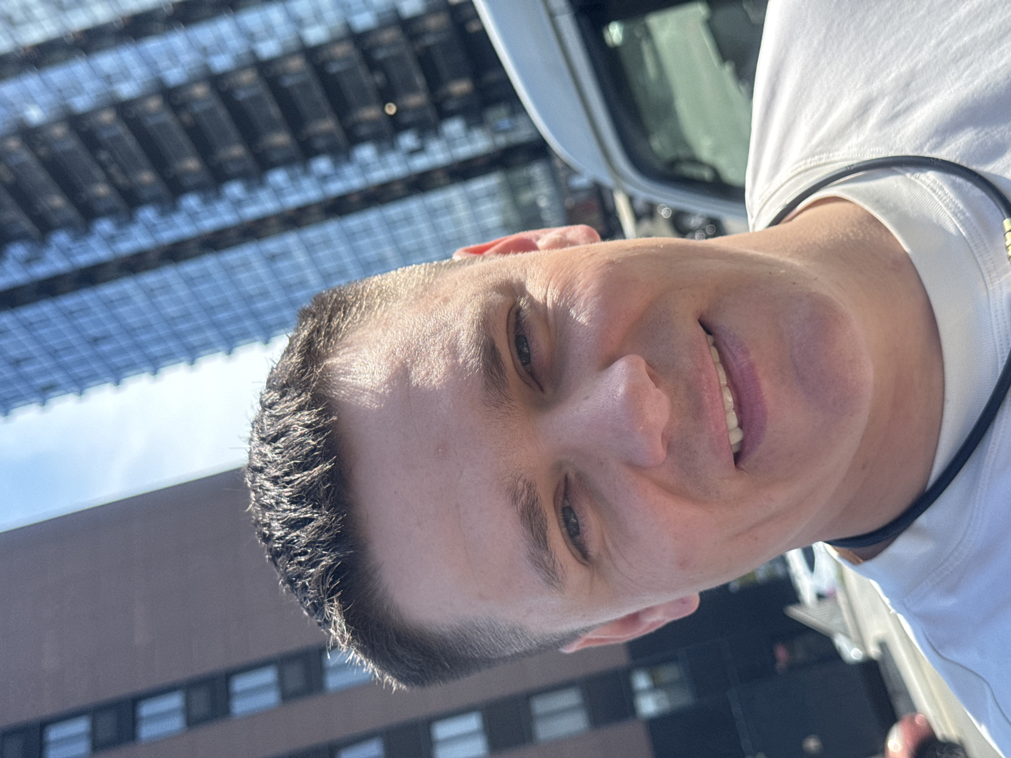 A man with short, dark hair and a fair complexion is shown in a close-up selfie. He is wearing a white t-shirt and a black necklace. His eyes are open and he is smiling, revealing his teeth. The background consists of modern buildings with glass facades and a clear blue sky. The scene appears to be set in a city, likely Vladivostok, Russia, given the architectural style of the buildings and the presence of vehicles. The sunlight suggests it is daytime.