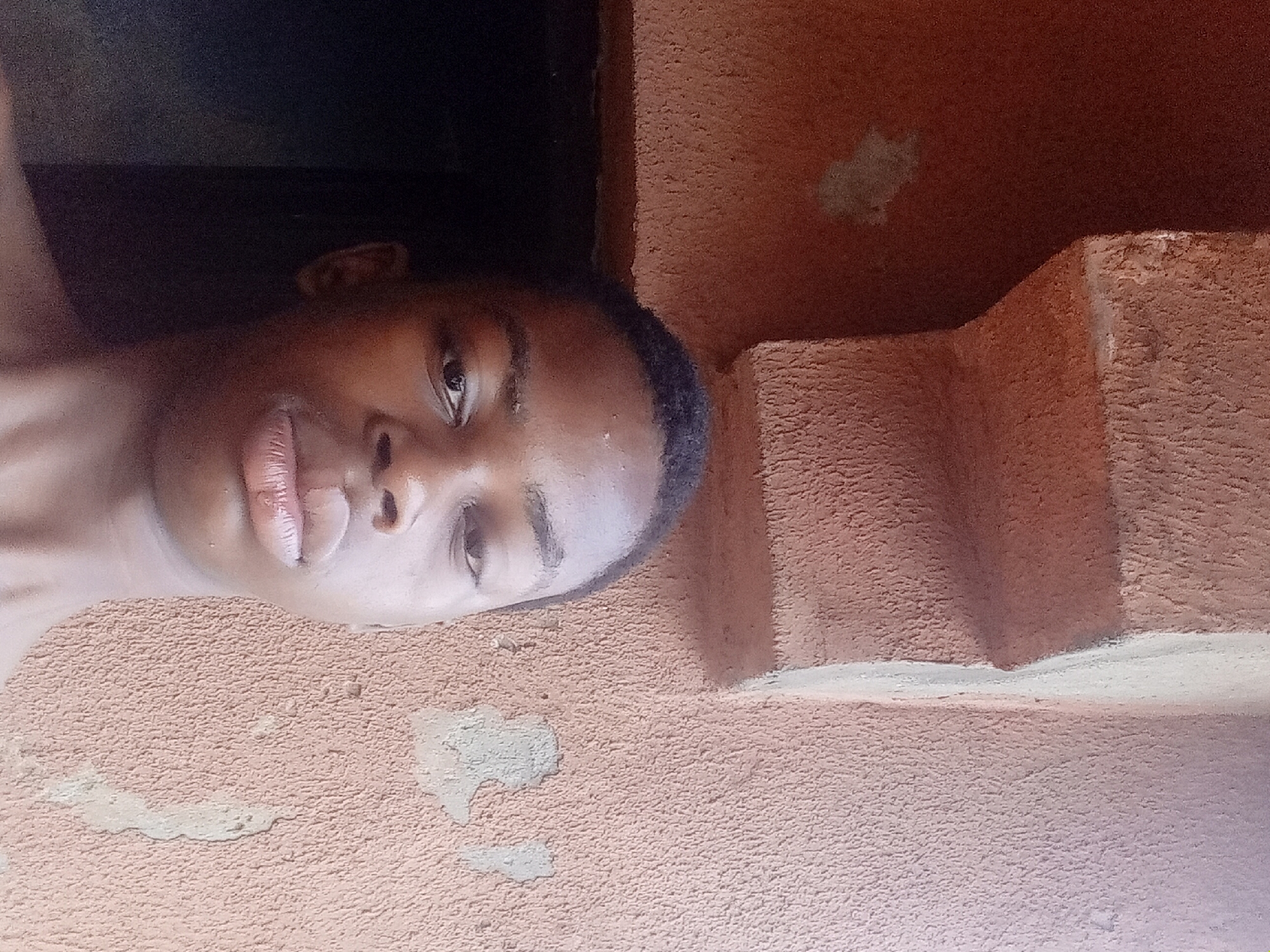 A young, dark-skinned person is pictured in a partially obstructed shot. They have short, dark hair. They are positioned against a textured wall with a rough, mottled surface. A dark, undefined space is visible behind them. The location is likely Nkpor, Nigeria, based on user input. There is some wall structure or steps next to the person.