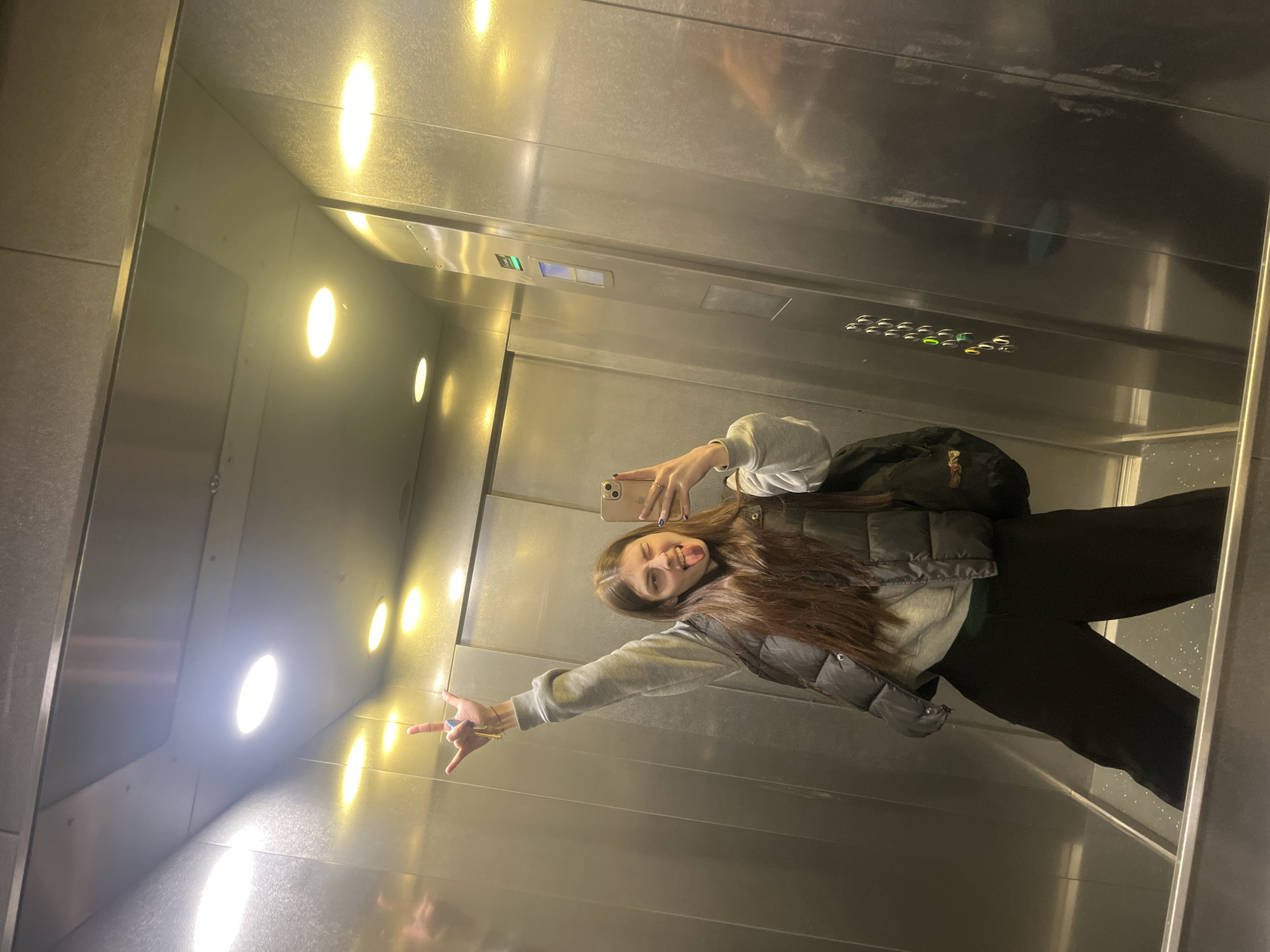 A woman is taking a selfie in an elevator. The elevator's interior is stainless steel with recessed lighting. The elevator's control panel is visible. The woman is wearing a gray sweater, a black vest, and black pants, and has a black backpack. She's holding a cell phone and making a hand gesture, with her tongue sticking out. The elevator appears to be located in Saint Petersburg, Russia.