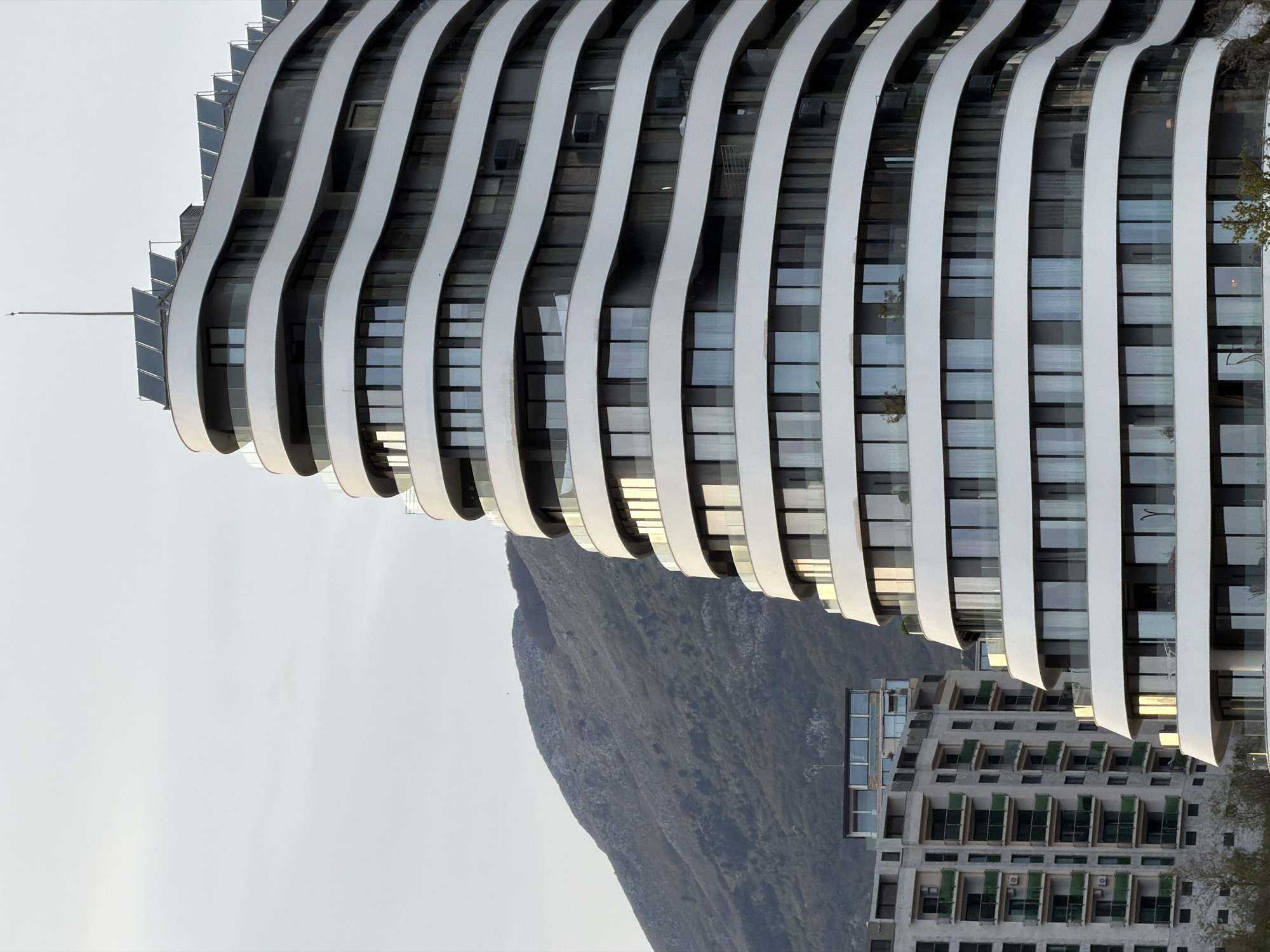 The image features a partial view of a modern architectural structure, likely a high-rise building, in Boreti, Montenegro. The building exhibits a distinctive design characterized by undulating, horizontal white bands running across its facade, creating a wave-like effect. Rectangular windows are visible between the bands, reflecting light. In the background, a mountain rises, partially obscured by the building. Another building with a more traditional, stepped facade is also visible near the bottom right corner. The sky is overcast, suggesting a dull or diffused lighting condition. There are no people visible. The scene depicts an architectural contrast between modern and older buildings, set against a mountainous backdrop.