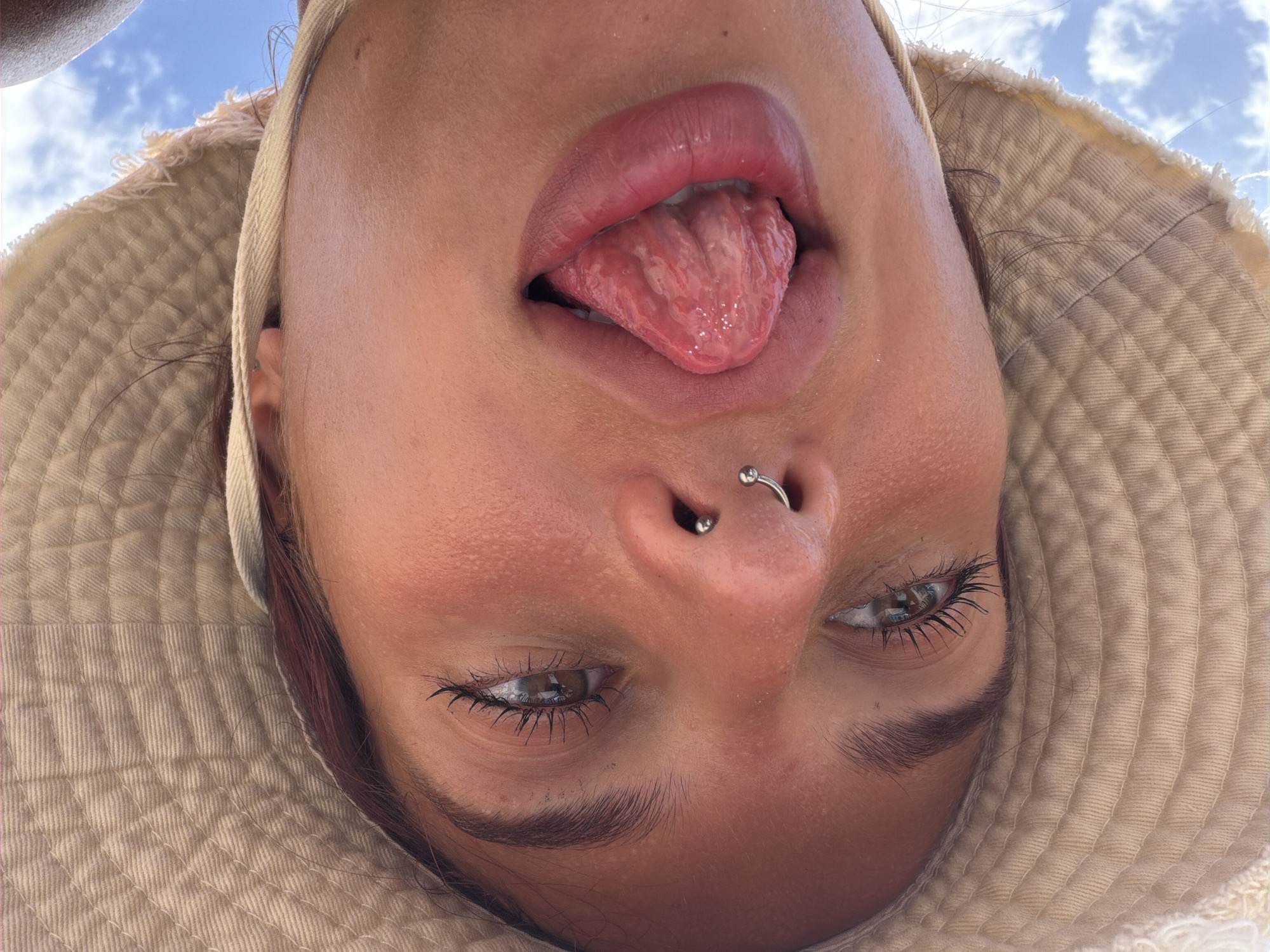 A low-angle, upside-down close-up shot shows a person wearing a tan bucket hat. The person's eyes are open and looking sideways. They have a septum piercing. Their mouth is open, and their tongue is sticking out. The tongue appears to be split or heavily textured. The background consists of a bright blue sky with scattered clouds. The image was taken in Arugam Bay, Sri Lanka.