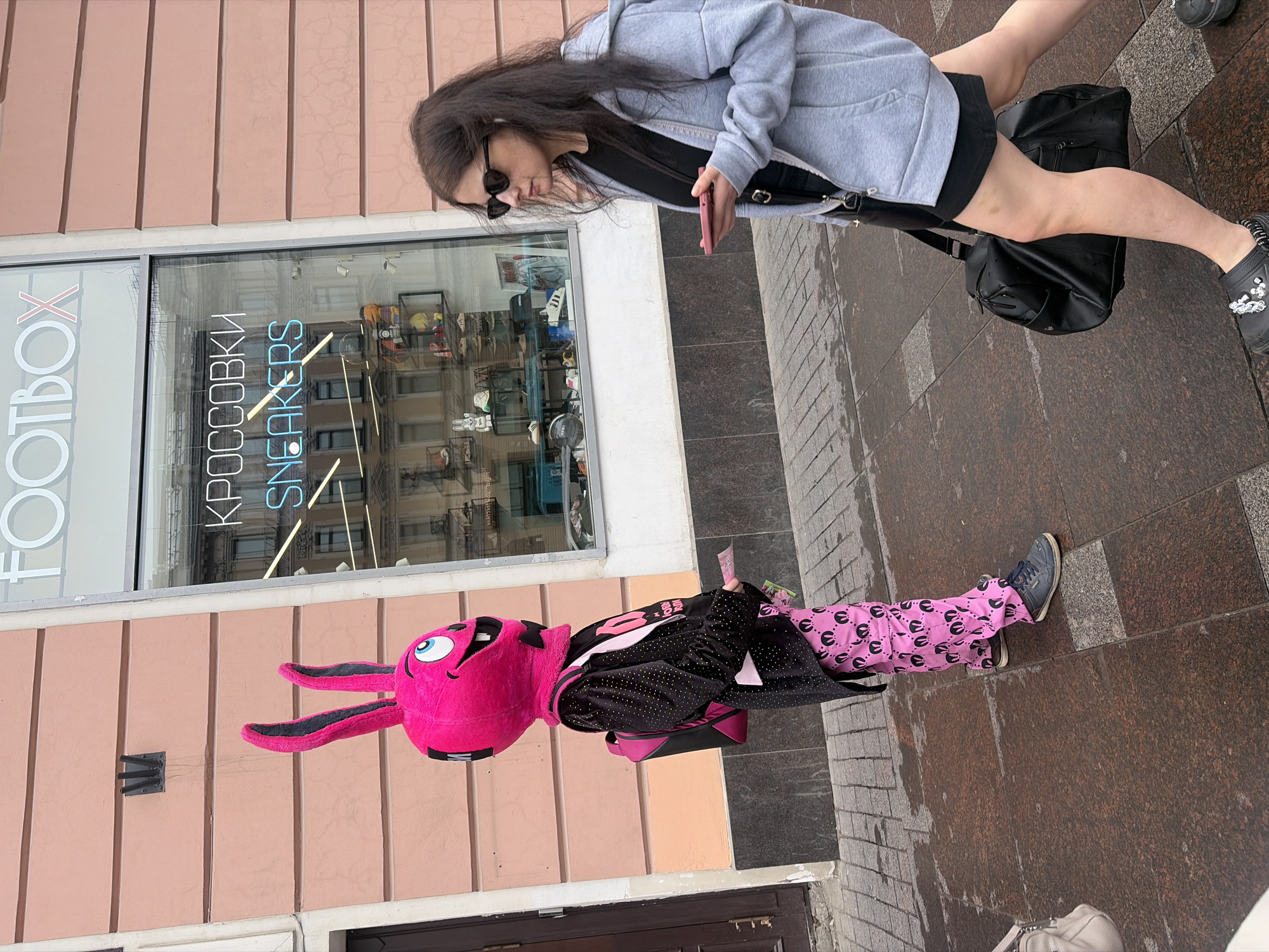 A person in a bright pink rabbit costume with large ears and blue eyes is standing on a sidewalk in front of a storefront. The costume has a black bow tie and is accessorized with a black jacket adorned with rhinestones and a pink backpack. The person is wearing pink pants with a black repeating pattern and blue sneakers. The storefront has a large window displaying shoes and a neon sign that reads "FOOTBOX" and "КРОССОВКИ SNEAKERS". To the right of the costumed person, a woman with dark hair and sunglasses is walking away, looking at her phone. She is wearing a grey hooded jacket over a black top and shorts, carrying a black duffel bag. The sidewalk appears wet, indicating recent rain. The building facade is painted a light pink hue. This scene is located on Nevsky Prospekt in Saint Petersburg, Russia.