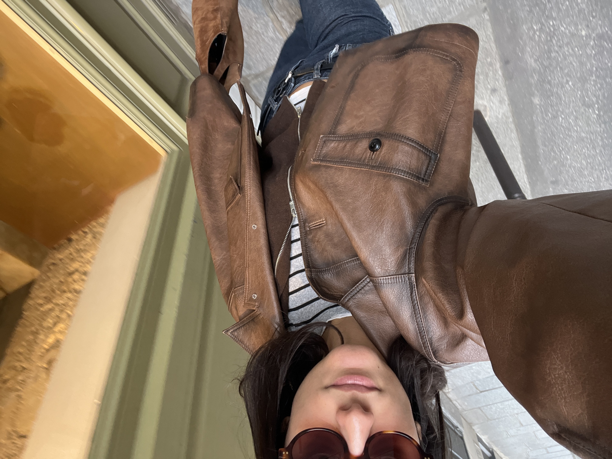 A high-angle selfie captures a young woman standing on a grey cobblestone pavement in what appears to be Paris, France. She wears dark, tortoiseshell-framed sunglasses that obscure her eyes, and her short, dark hair frames her face. Her attire consists of a brown leather jacket with a distressed finish, featuring a collar and a visible buttoned chest pocket with a black button. Underneath, a brown full-zip garment is partially open, revealing a white and grey horizontally striped top. She wears dark blue jeans with a belt and holds a brown leather-like bag in her right hand. To her right, a green multi-paneled wooden building facade with white trim is visible, featuring a rectangular window reflecting an indistinct, warm-toned interior, possibly with a wooden staircase. A section of a white wall adjoins the green facade. A thin black vertical pole is partially visible on the left side of the frame.