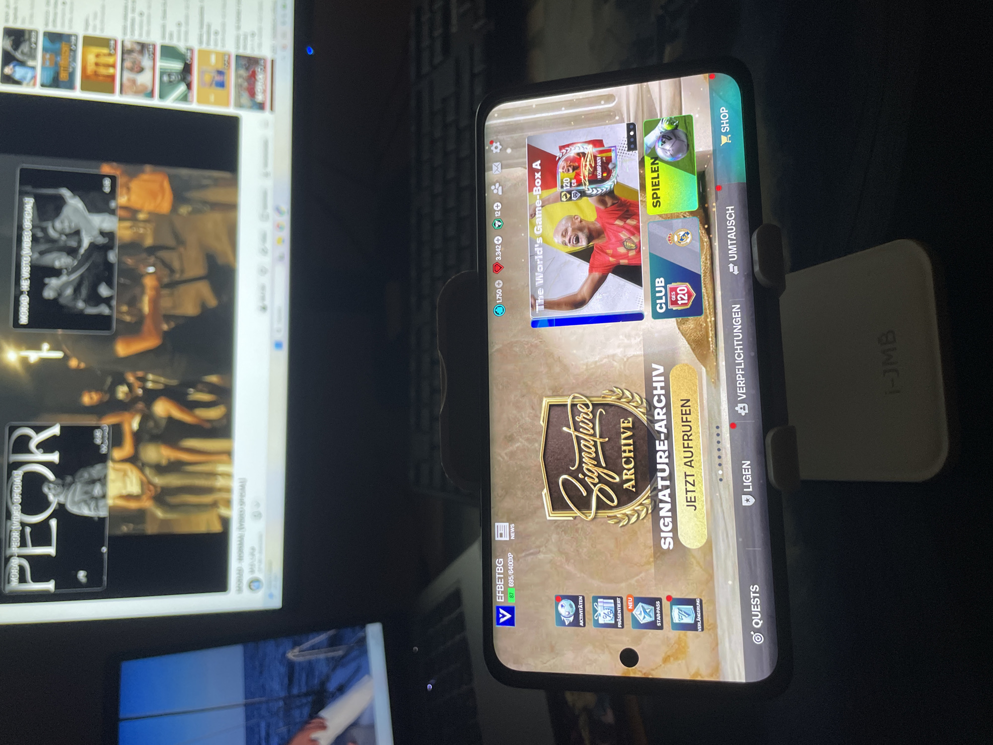 The image shows a person playing a mobile game on a smartphone, which is propped up on a stand. The game appears to be a soccer-themed card collecting game. In the background, a computer monitor displays a YouTube video. The setting is indoors, with a keyboard visible on the desk. The lighting suggests it is nighttime. There are no specific geographical markers to indicate a city or location.