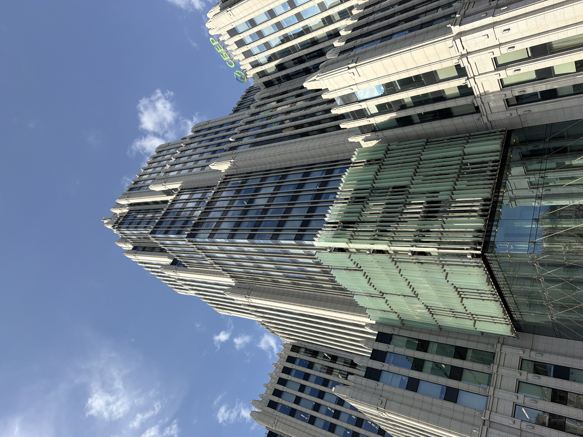 The image shows the "Sberbank City" skyscraper in Moscow, Russia. The building is angled upwards towards the sky, revealing its architectural details. It features a combination of glass and stone, with horizontal lines forming distinct layers. The lower section includes a green-tinted glass design. The sky is blue with scattered white clouds. The angle suggests the image was taken from ground level.