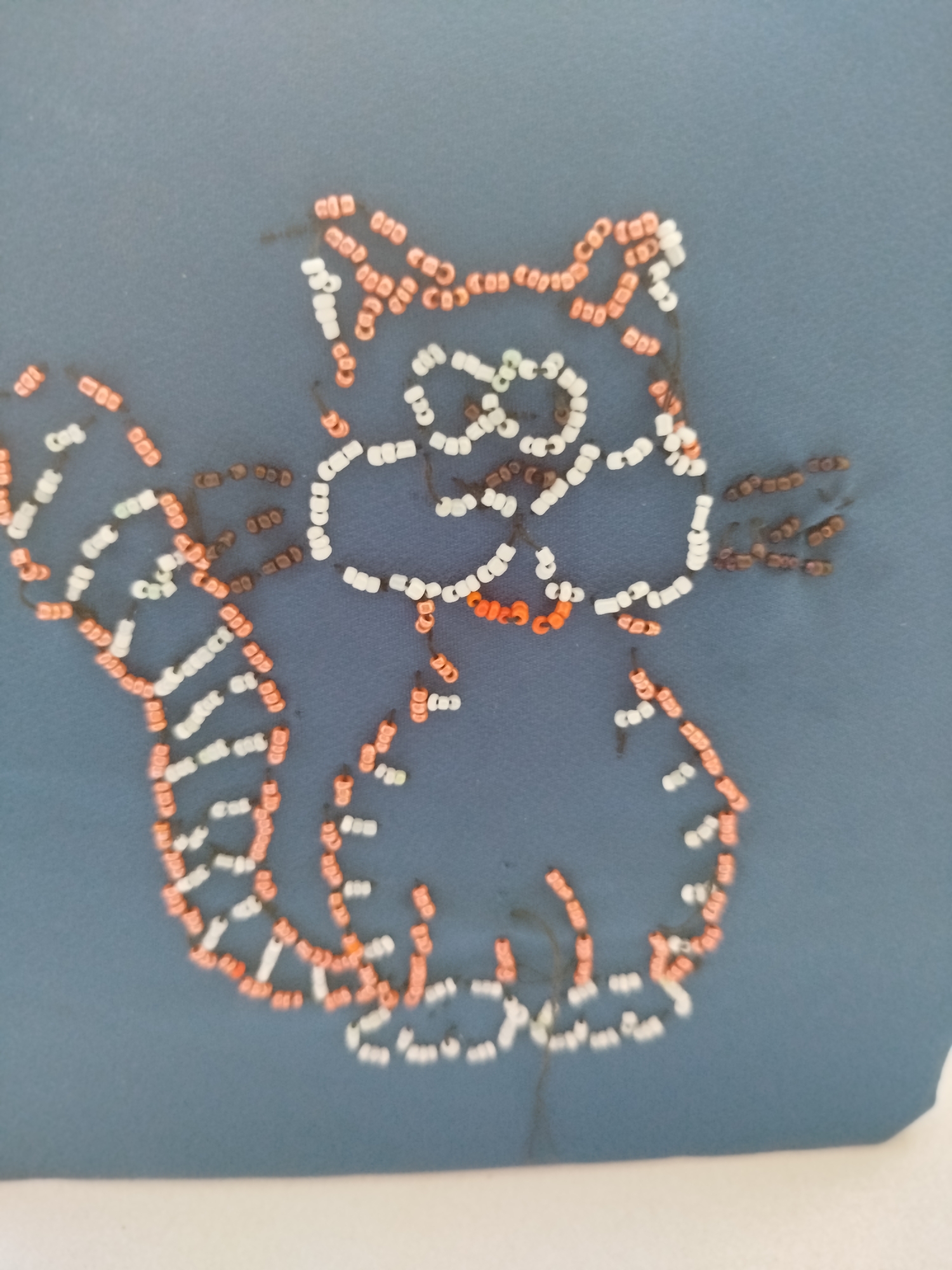 This image displays a beaded craft of a cat on a dark blue fabric. The cat is outlined and filled with small white, orange, and brown beads. The craft appears to be a decorative element, possibly part of clothing or an accessory. There is no discernible information about the location in Крывасельскі сельскі Савет, Belarus.