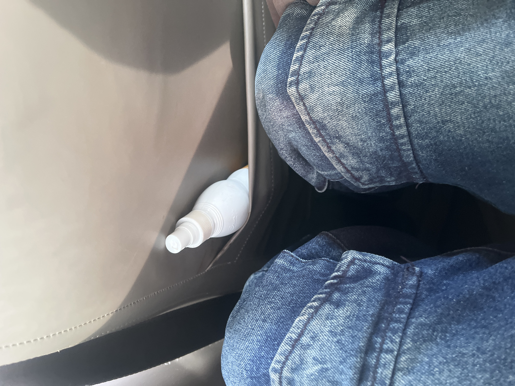 The image shows a close-up view of a person's legs wearing blue denim jeans. The person is seated, and the jeans are folded or creased, suggesting they are in a confined space. To the left of the jeans, there is a pocket within a bag or on a piece of furniture, and a small white spray bottle is visible inside this pocket. The bottle has a nozzle and a cap. The material of the bag or furniture appears to be a light brown or beige, possibly leather or a similar synthetic material. The lighting suggests it is daytime, with natural light illuminating the scene. There are no visible landmarks or distinct cues to determine the specific location within Jalingo, Nigeria, beyond what is implied by the provided context. The image is a still shot, and there are no explicit indications of emotions or a specific event taking place. The focus is on the texture of the denim and the white spray bottle against the neutral background.