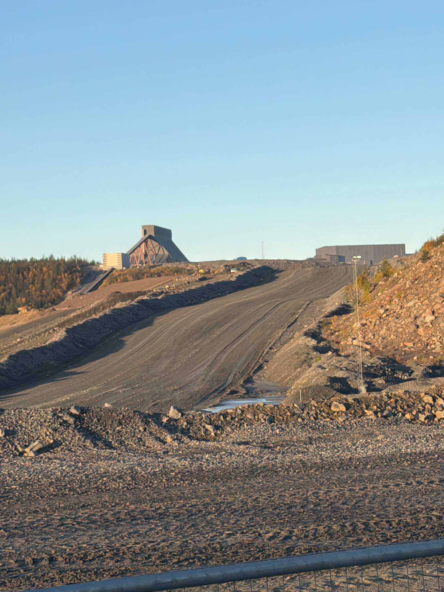 An industrial landscape in Gällivare, Sweden, features a large mine or processing plant. In the background, a distinctive grey, angular structure with red scaffolding is situated on a hillside covered with sparse trees showing autumn colors. Adjacent to it, a long, grey, box-like building is visible. The foreground is dominated by vast piles of dark grey, loose rock and gravel, indicative of mining or construction activity. A paved, winding road or path cuts through the mounds of rubble, leading uphill towards the structures. There are no people visible in the image. The sky is clear and blue, suggesting daytime. The overall scene depicts an active or recently active industrial site with significant earthmoving operations.