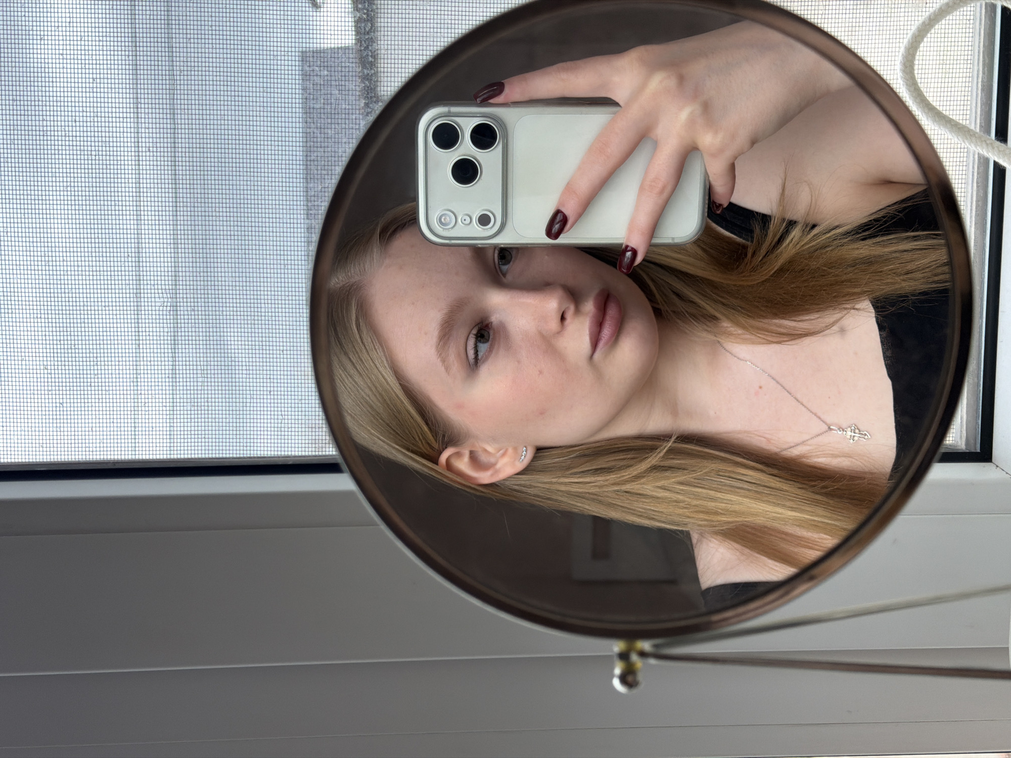 A person is taking a selfie in a round mirror. The person has light skin, light brown or blonde hair, and is wearing a necklace with a cross pendant. They are holding a white smartphone with multiple camera lenses in their right hand, partially obscuring their face. The nails on the hand are painted a dark color.
The mirror is positioned in front of a window, which has a screen. The background visible through the window appears to be an indistinct outdoor scene. The window frame and a windowsill are visible around the mirror. A white cord is also seen near the top right corner of the mirror. It is not possible to determine the location as Rossiyskiy, Russia or any specific city location from the provided image.