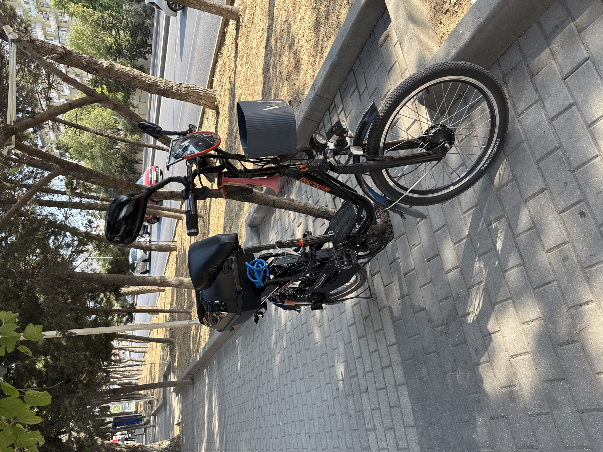 A black electric bicycle is parked on a paved sidewalk next to a tree-lined street. The bicycle has a storage box on the back, a blue cable lock, a red horn, and a dark grey basket attached to the handlebars. In the background, cars are driving on the street, and apartment buildings are visible. The scene appears to be in Baku, Azerbaijan, given the architectural style of the buildings and the presence of street trees commonly found in the city. The lighting suggests it is daytime, likely late afternoon due to the long shadows cast by the trees.
