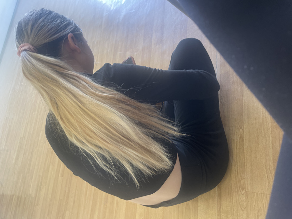 A person, likely female, is seated on a light-colored wooden laminate floor, viewed from an elevated rear-right angle. Her long, blonde hair is secured in a high ponytail with a light pink scrunchie. She is dressed in a form-fitting black long-sleeved top that exposes a portion of her lower back, and black leggings. Her head is bowed, directed downwards, and she is holding a smartphone, partially visible in her right hand and equipped with a reddish-pink case. The environment is an indoor space with bright light reflecting off the polished floor. A dark, out-of-focus object is present in the lower right and left foreground. The specific location within Sofia, Bulgaria, cannot be determined from the visual information provided; the setting is a generic indoor space.