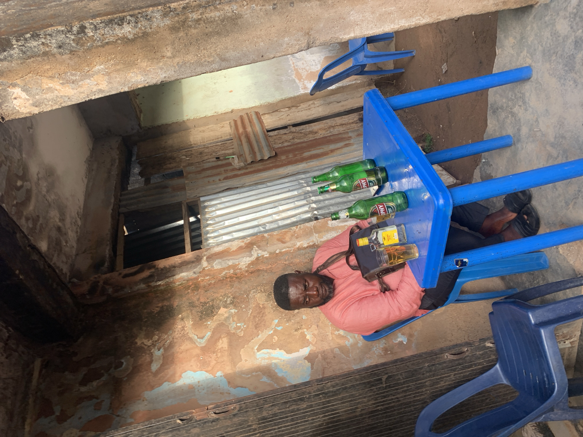 A man wearing a pink shirt and dark pants is lying on a blue plastic table with bottles of alcohol on it. He is outdoors, seated near dilapidated walls, some with peeling paint and corrugated metal. There are several blue plastic chairs scattered around. The location appears to be in the city of Jos, Nigeria.