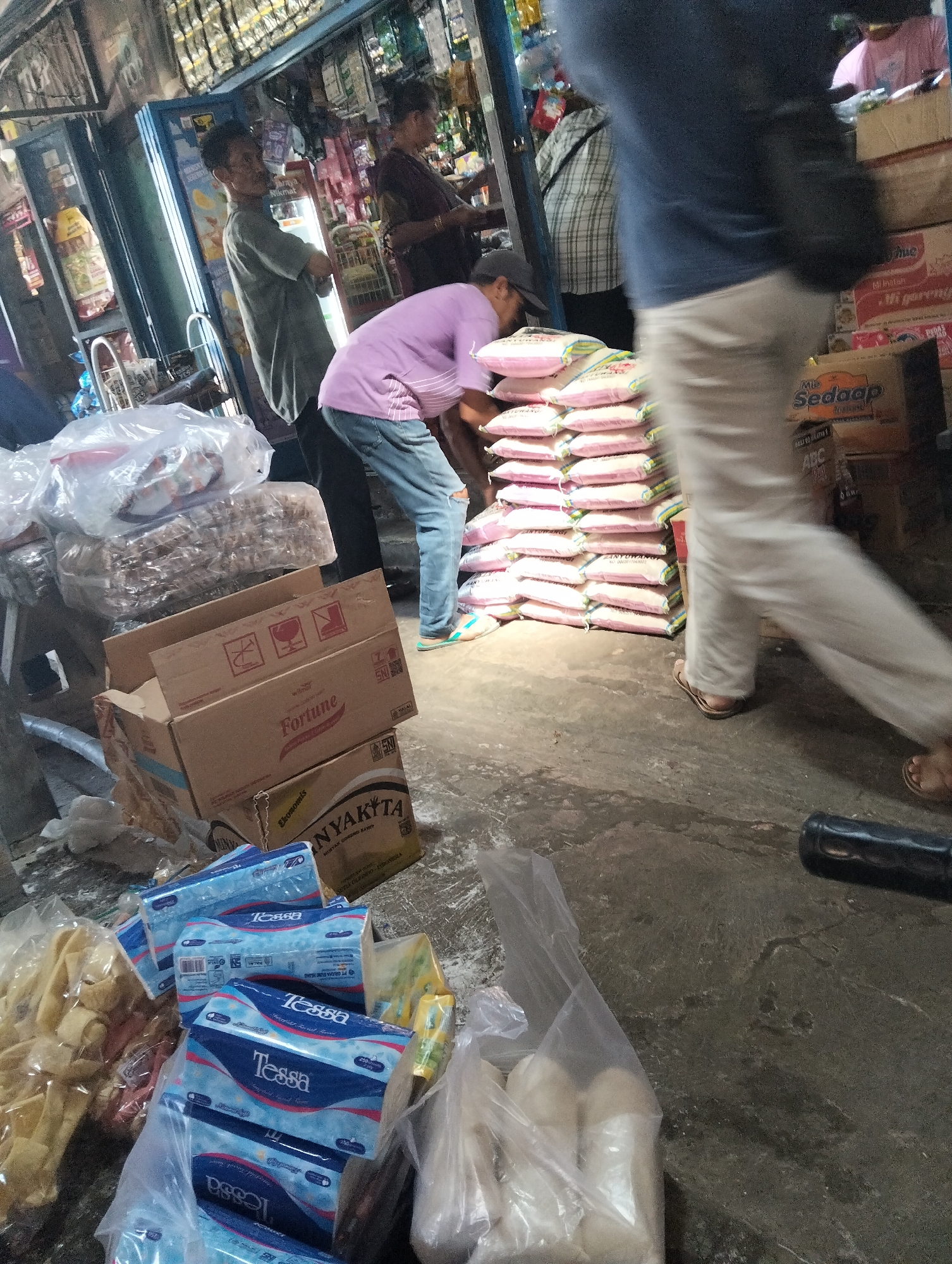 The image depicts a scene inside a dimly lit store, possibly a market stall, located in Muncar, Indonesia. The ground is made of rough concrete, and the walls appear to be constructed of various materials, including what looks like corrugated metal or plastic sheeting. Stacks of goods, predominantly packaged food items like rice and what appear to be tissue boxes (branded "Tessa"), are visible. Several people are present. In the foreground, a person wearing blue jeans and a purple shirt is seen bending down, interacting with a stack of rice bags. To the left, a person in a grey shirt stands with arms crossed, looking towards the center. Further back, another individual in a dark top is partially visible, and a person in a plaid shirt is also present. The overall impression is one of commercial activity in a somewhat cluttered and modest retail environment.