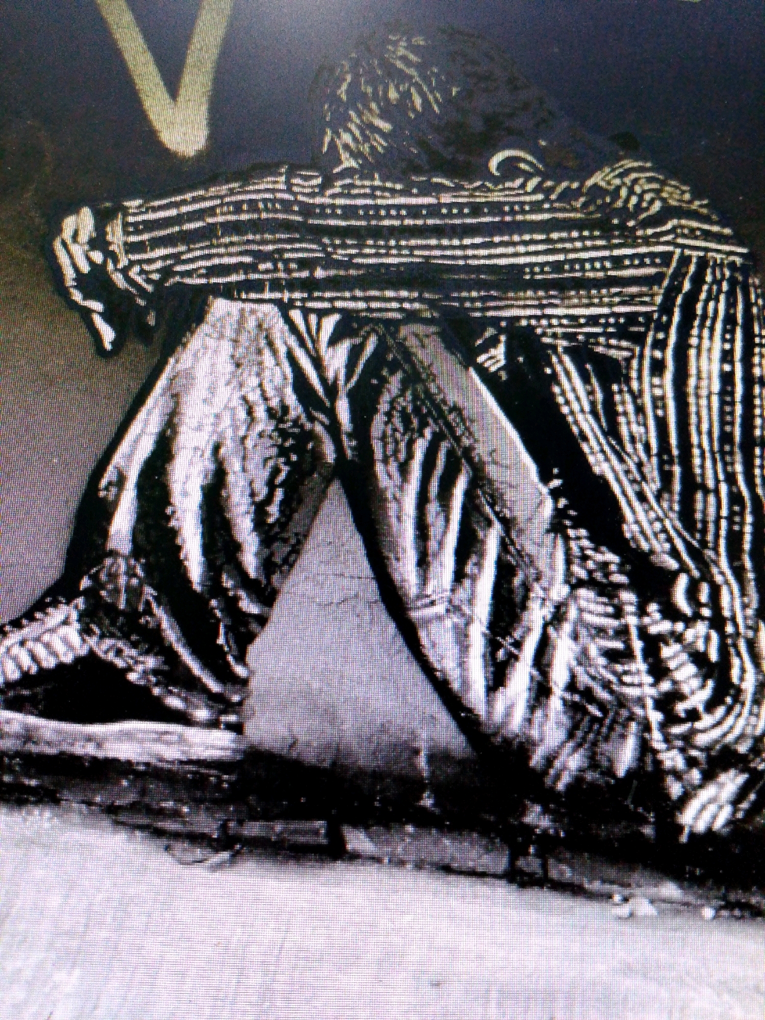 A person is depicted sitting on the ground with their knees drawn up to their chest, their head bowed, and their arms wrapped around their legs. They are wearing a horizontally striped shirt and pants. The background is dark with a bright, V-shaped light source above and to the left of the person. The image appears to be a photograph with a visible pixelated texture, suggesting it may have been taken with a low-resolution camera or a digital zoom. No specific details of the city of Glonggong, Indonesia, are visible in the image.