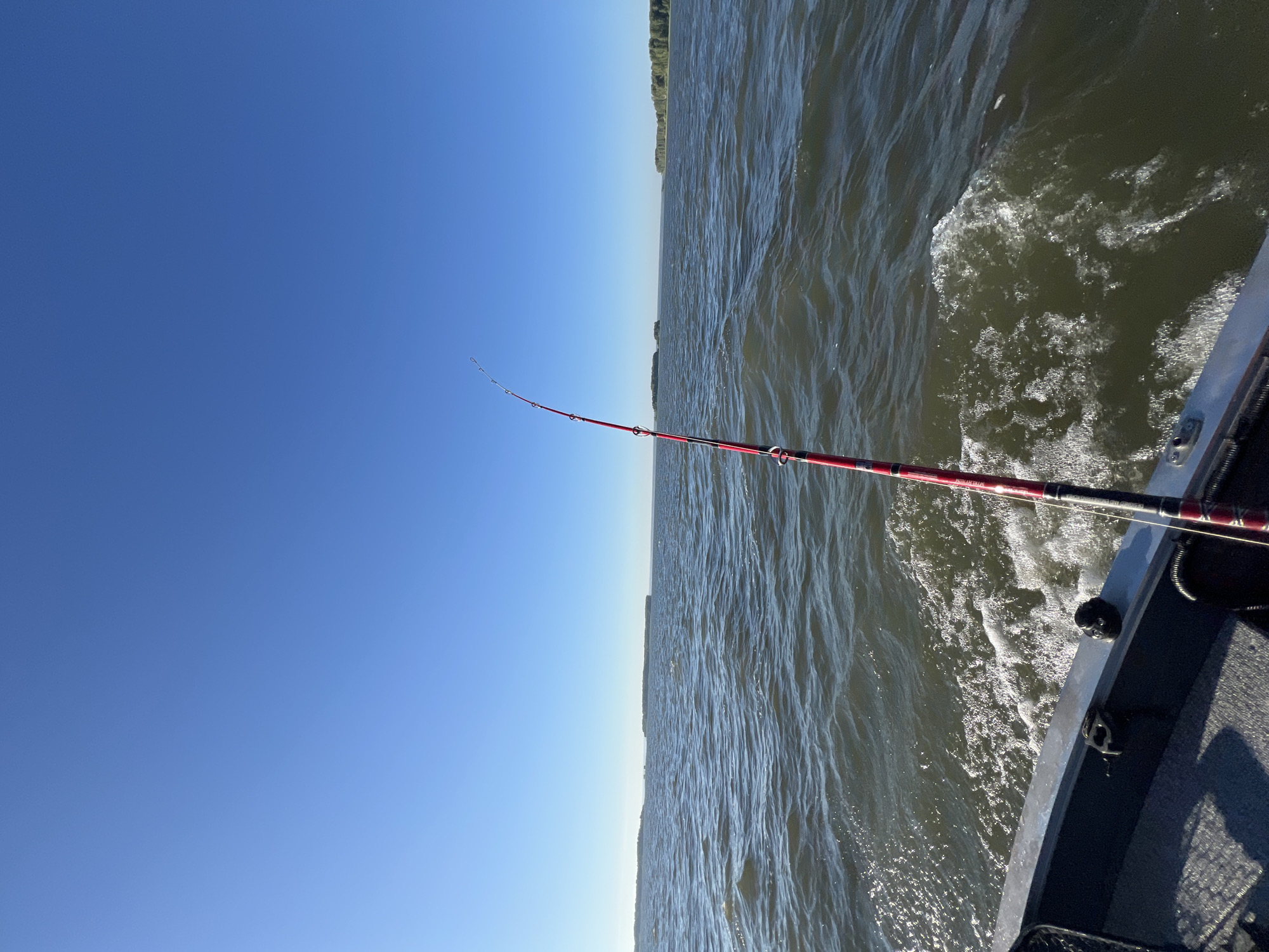 An eye-level, medium shot captures a clear blue sky over a large body of water. The foreground is dominated by the edge of a gray boat, with a fishing rod, red and black, extending diagonally upwards. The water is choppy, with white foam trailing behind the boat, indicating movement. In the distance, several tree-covered islands or peninsulas dot the horizon. The scene appears to be a serene fishing excursion on a sunny day. No people are visible in the frame. The location is specified as Novopichugovo, Russia.