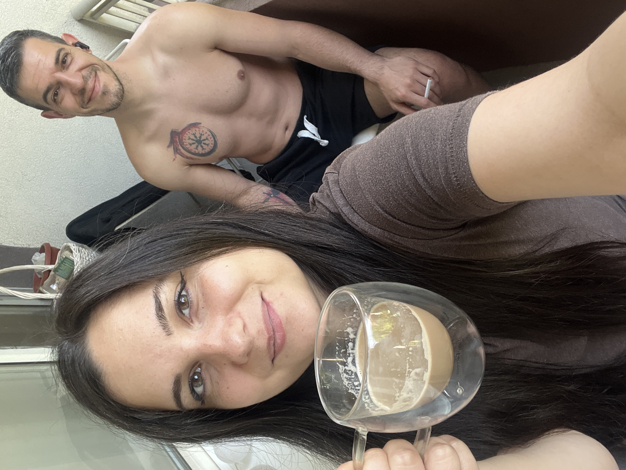 This is a selfie taken by a woman holding a double-walled glass mug of coffee. She is wearing a brown t-shirt and has long dark hair. Beside her, a man with short brown hair and a beard, wearing black shorts, is seated. He has a tattoo of a circular design with a wing on his chest and a smaller tattoo on his upper arm. He is smiling and appears to be wearing a wireless earbud. His right hand is resting on his lap, and he is holding a cigarette. The background suggests an outdoor setting, possibly a balcony, with a railing and a wall textured with a rough plaster finish. A woven basket with plants and a terracotta pot is visible behind the woman. The lighting indicates it is daytime.