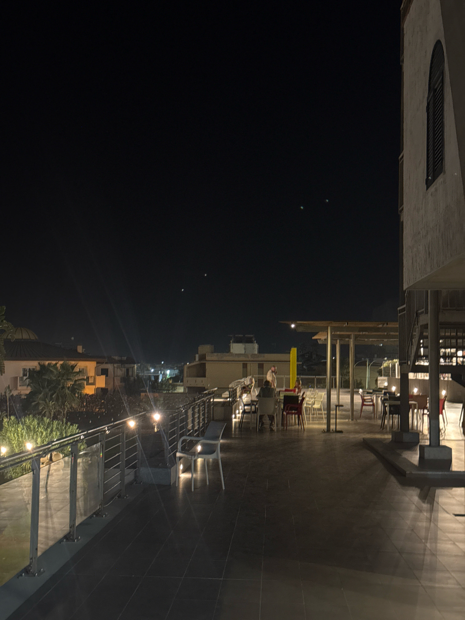 The image depicts an outdoor terrace at night, possibly a rooftop restaurant or bar, located in Capaci, Italy. The scene is dimly lit by string lights and ambient light from the surrounding city and buildings. In the foreground, a dark tiled floor is visible, bordered by a metal and glass railing. Several tables and chairs are arranged on the terrace, some occupied by patrons. A few people are seated at tables, engaged in conversation or dining. A person is standing near a yellow structure in the background, talking to another individual seated at a table. The sky is dark, with a few faint lights that could be stars or distant lights. The architecture suggests a Mediterranean style with a building on the right featuring an arched window. The overall impression is one of a relaxed evening gathering.