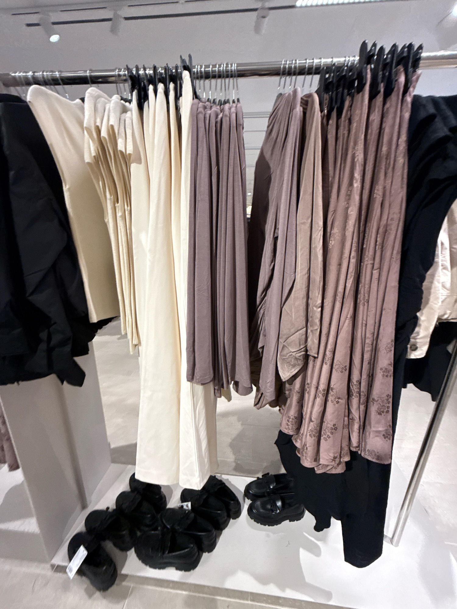 The image shows a clothing store display in Tallinn, Estonia. A rack of clothing, including black jackets, cream-colored pants, and mauve dresses, is visible. Several pairs of black loafers are arranged on the floor in front of the clothing rack. The background consists of white walls and a white floor, with metal shelving and lighting fixtures typical of a retail environment. No people are present in the image.