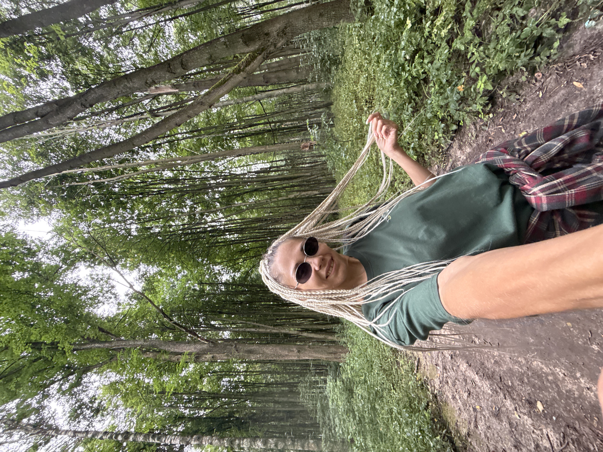 A woman with blonde braided hair and sunglasses is taking a selfie while walking on a dirt path in a forest. She is wearing a green t-shirt and has a plaid shirt tied around her waist. The forest is dense with tall trees and green foliage. The location is the city of Dumino, Russia.