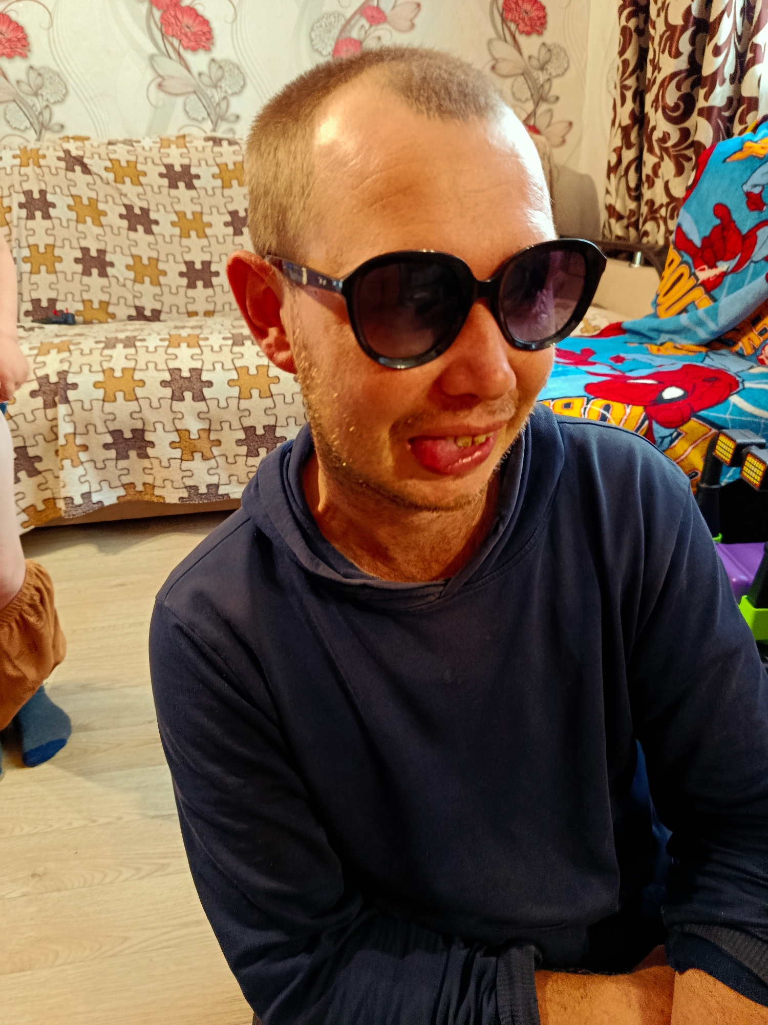 A medium shot captures a man wearing dark sunglasses and a dark blue hoodie, seated on a light-colored wooden floor. His tongue is slightly extended, and he has a light stubble. In the background, a patterned couch with a puzzle piece design is visible, along with a wall with floral wallpaper and patterned curtains. A colorful blanket, possibly featuring cartoon characters like Spider-Man, lies on a surface behind the man. The overall environment suggests an indoor domestic setting.