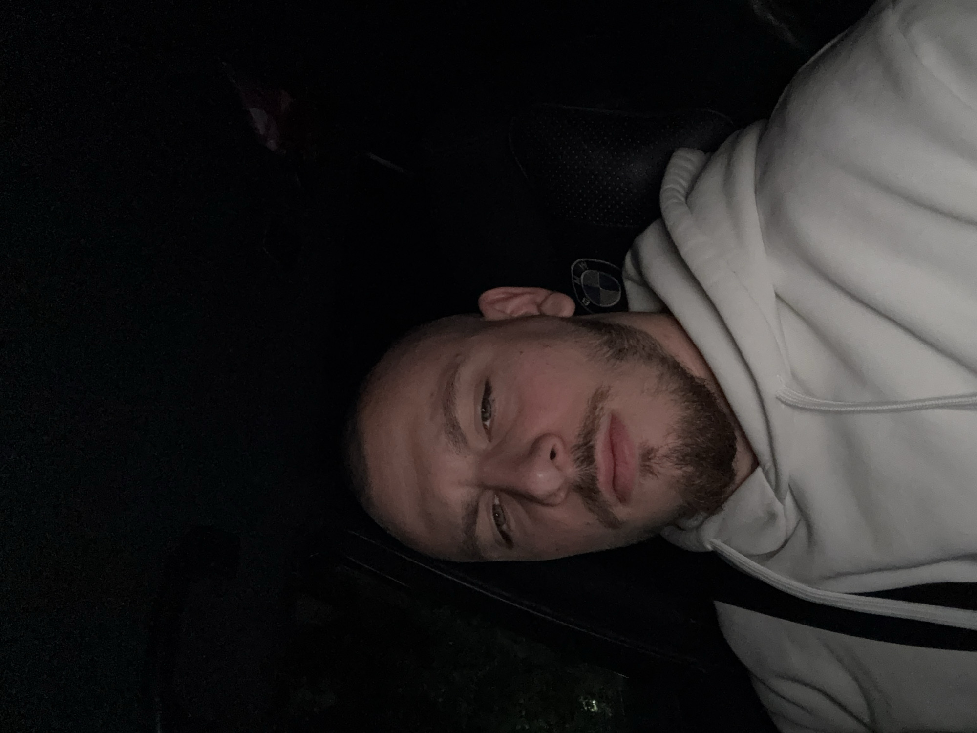 The image shows a person with a shaved head, a beard, and wearing a white hoodie with a black stripe. The person is lying down in what appears to be the interior of a car, based on the presence of a headrest and a possible car window. The background is predominantly dark, suggesting a low-light environment, potentially at night. There is a BMW logo visible, seemingly on the car's interior. The location cannot be determined to be in the city of Stara Zagora, Bulgaria, without additional contextual information from the image.