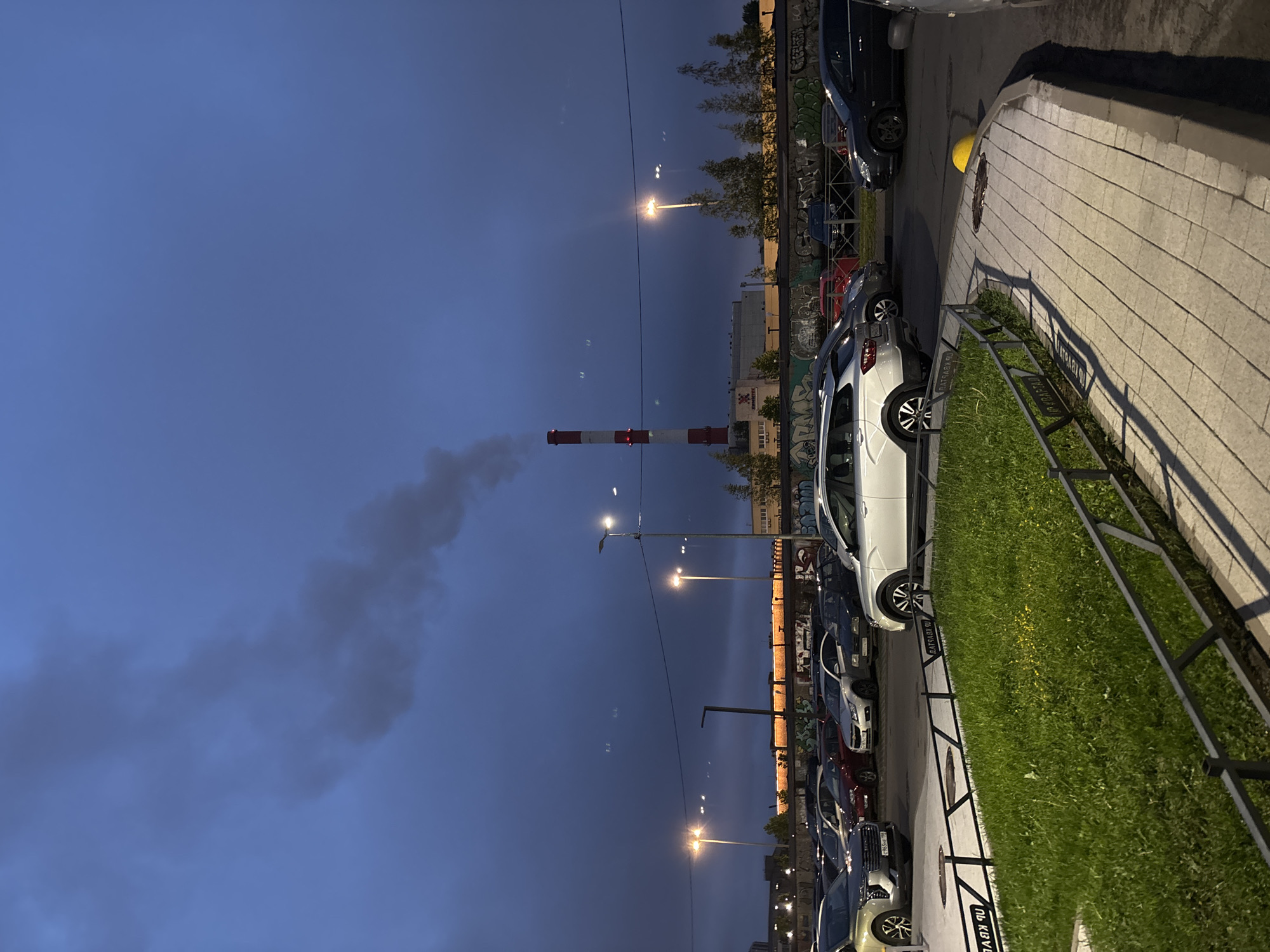The image depicts an urban scene at dusk, featuring a row of parked cars along a street with a curb and grass area. Streetlights illuminate the scene, and a tall industrial chimney with red and white stripes is visible in the background, emitting smoke. A building or structure is situated near the chimney, and graffiti decorates a wall alongside the parked cars. The sky is dark, indicating twilight or early evening.