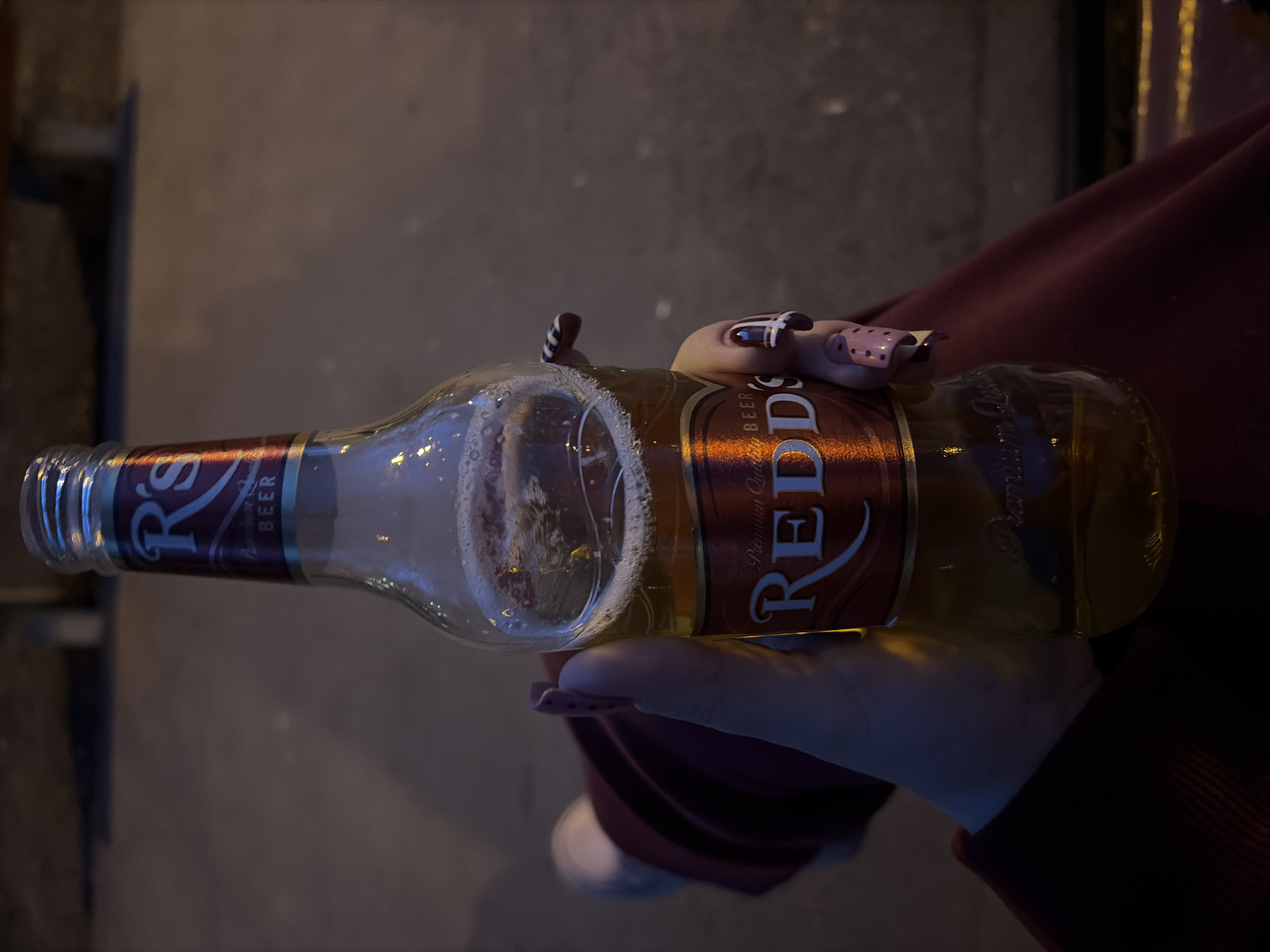An eye-level shot captures a hand holding a bottle of "Redd's" beer in a dimly lit setting. The bottle, made of clear glass, is about half full with a golden-colored liquid and shows some foam at the top. The label is predominantly red and brown, featuring the brand name "REDD'S" in large, stylized white letters. Below it, smaller text reads "Premium Quality" and "BEER."
The hand holding the bottle appears to be a woman's, with distinctive, elaborately painted fingernails. Some nails are pink with darker spots, while others are dark with white stripes or patterns. The fingers are wrapped around the bottle, suggesting a firm but relaxed grip. A sleeve of dark red or burgundy clothing is visible around the wrist and arm.
The background is dark and indistinct, with soft, out-of-focus elements. On the left, a dark vertical object, possibly part of a structure or furniture, can be seen. On the right, hints of draped fabric in a similar dark red color as the clothing are visible. The overall lighting is low, suggesting it might be evening or night, or an indoor setting with subdued illumination. The ground or surface beneath the bottle is a muted, dark gray or brown, possibly concrete or asphalt. No specific location cues for Perm, Russia are visible within the image itself.
