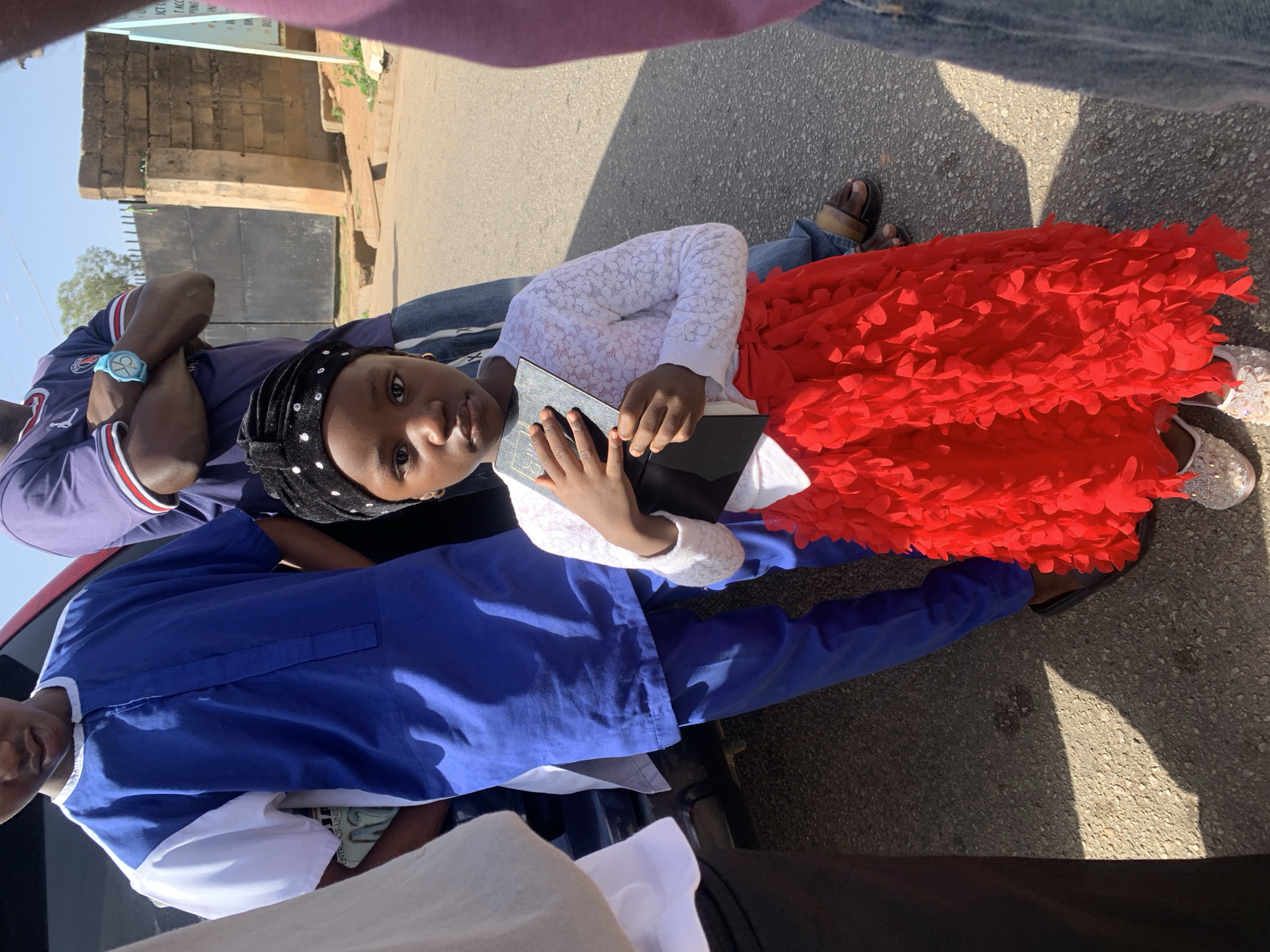 A young girl in a white lace-patterned top and a red floral skirt, wearing a black head covering, holds a dark-covered book. She is surrounded by people, including one wearing a blue and white shirt and another in a blue top and matching trousers. The setting appears to be an outdoor street in Jos, Nigeria. There is a building in the background with a black gate and a sign above it. The pavement is grey.
