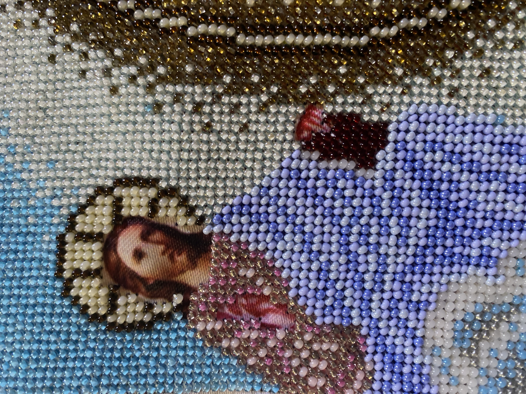 This is a close-up, high-angle, medium shot of a beaded tapestry depicting Jesus. The tapestry features a religious image with Jesus' face in the upper left quadrant, surrounded by a beaded halo. His robes are depicted with various colors of beads, including blues, purples, pinks, and white. A textured, possibly metallic, element is visible on the right side of the image, also made of beads, which may represent a tree or religious artifact. The background is a mosaic of light blue beads. The tapestry appears to be a detail from a larger religious artwork or religious garment. There is no visible indication of Zgrade, Montenegro, or any urban environment.