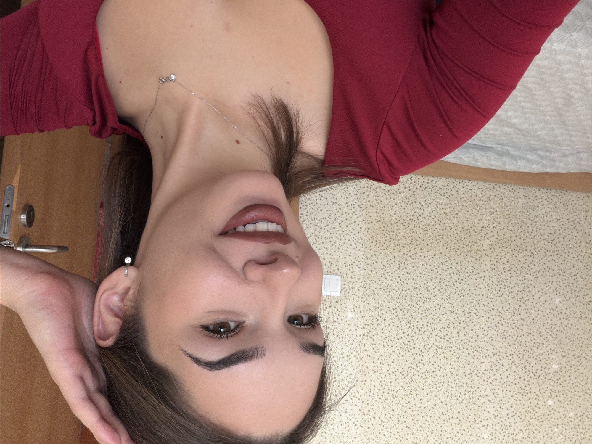The image displays a head and shoulders shot of a female subject, centrally positioned and looking directly at the viewer with a smile that reveals her teeth. The subject has light-toned skin, brown eyes, and dark brown hair, with prominent, well-defined eyebrows. She is wearing dark brown lipstick. Her right hand is raised, with fingers resting near the right side of her head and ear. She wears a silver-colored hoop earring with a small clear stone in her right ear and a silver-colored necklace with a small, clear-stoned pendant on her chest. Her attire consists of a red long-sleeved top.
The background consists of an interior setting. To the left and upper sections, a light beige wall with a speckled pattern of darker dots is visible. A white electrical outlet or switch plate with two rectangular buttons is mounted on this wall to the left of the subject's head. Below this and behind the subject's left shoulder, a segment of a wooden headboard or bed frame is present, above which lies light-colored, possibly greyish-white, patterned bedding. To the far right of the frame, a vertical section of a light brown wooden door is visible, equipped with a silver-colored door handle and lock mechanism. The image provides no discernible visual information to confirm the location as Sofia, Bulgaria.