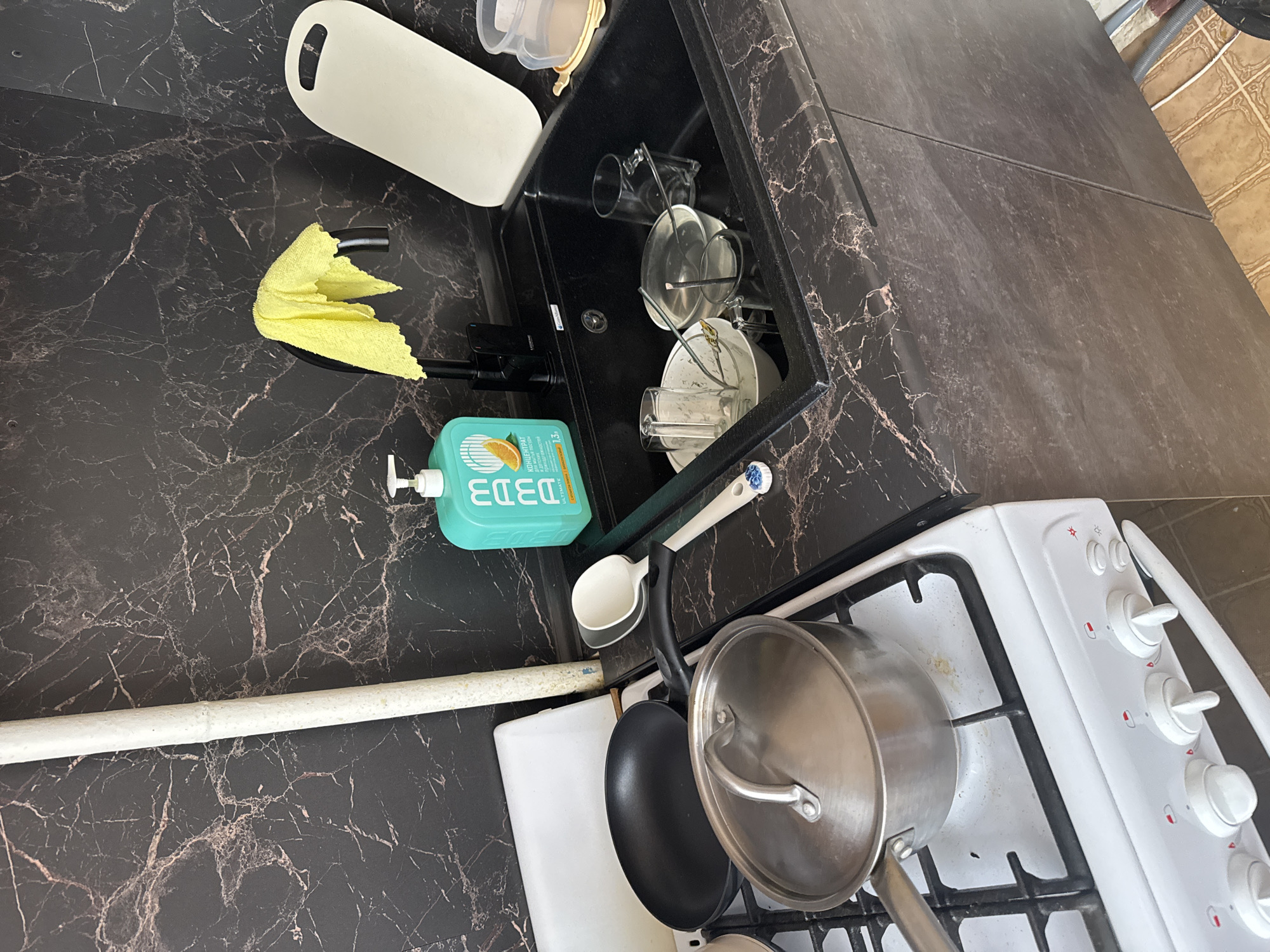 The image depicts a kitchen scene, likely indoors.
**Objects:**
* A black countertop with a marble pattern
* A sink with dirty dishes inside
* A white cutting board
* A yellow cleaning cloth
* A bottle of dish soap
* A cooking spoon and a dish brush
* A white gas stove with multiple burners and knobs
* Two pots and one pan on the stove
**Environment:**
* The floor is tiled with brown and tan tiles.
* The countertop and sink are situated next to the stove, indicating a typical kitchen layout.
Given the information regarding the city of Chaikovsky, Russia, it is possible the image originates from a kitchen in a home or apartment located in that city. There are no indicators as to a location in Chaikovsky.