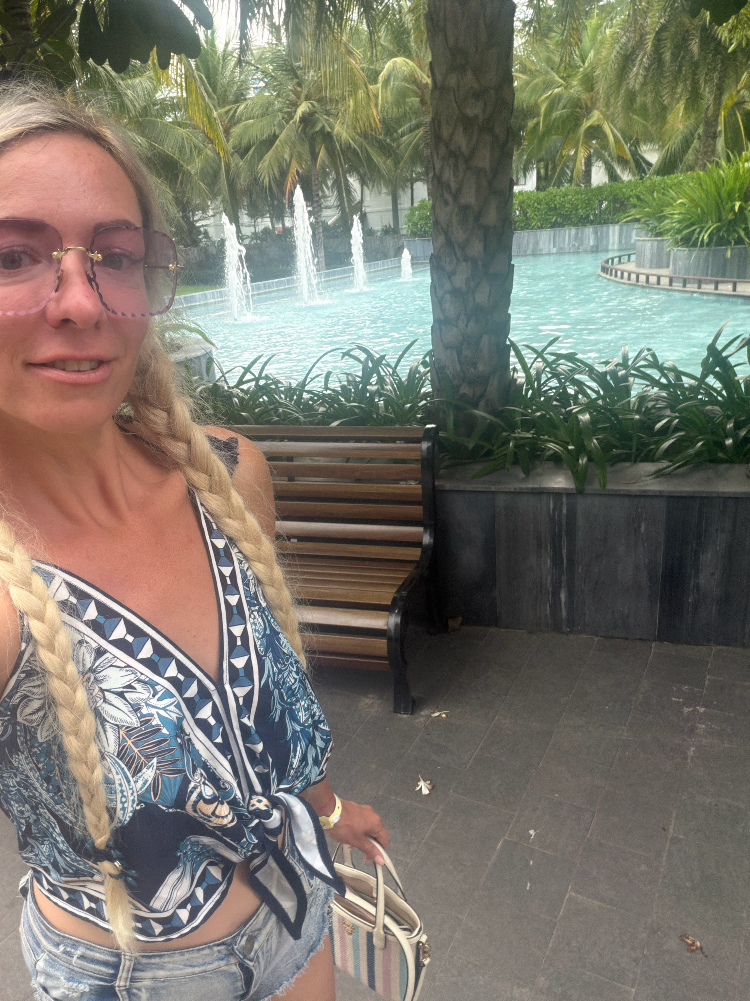 A Caucasian woman with blonde braided hair and pink-rimmed glasses is taking a selfie in an outdoor resort environment. She is wearing a blue and white patterned tie-front top and denim shorts. She is holding a white and striped handbag. Behind her, there is a swimming pool with fountains, palm trees, and lush greenery. The ground is paved with large, dark gray square tiles, and there is a wooden bench to her right. The scene appears to be in Cam Lâm, Vietnam, suggesting a tropical resort setting.