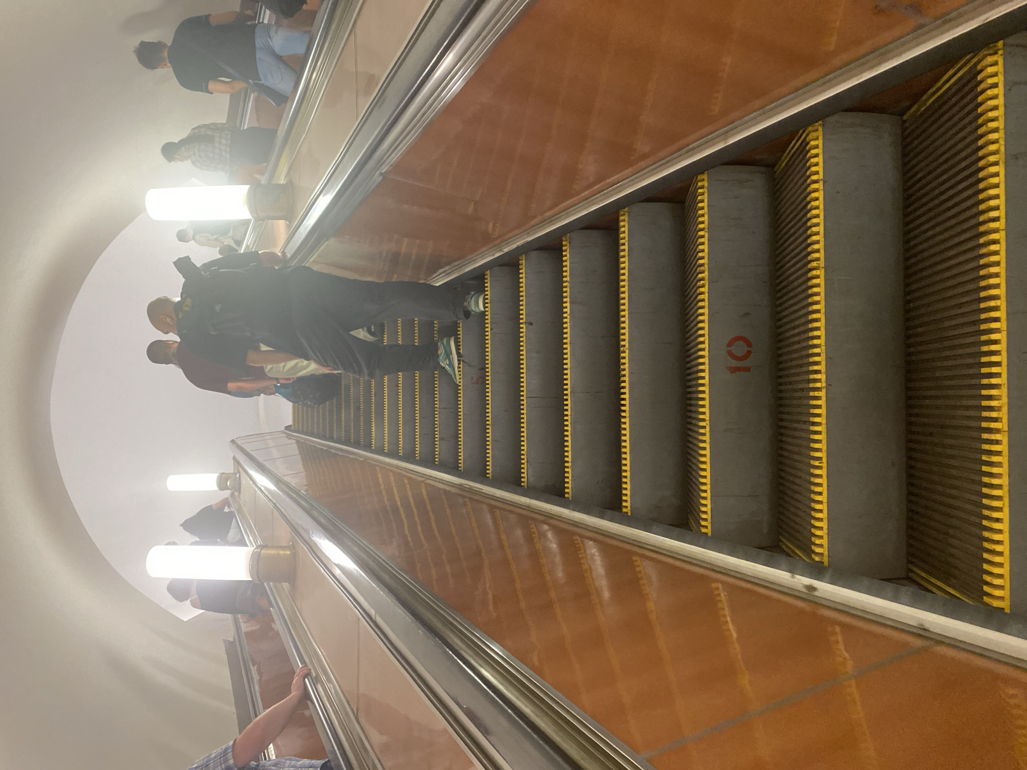 The image shows an upward-moving escalator in a Moscow Metro station, Russia. Several people are riding the escalator. The escalator steps are gray with yellow safety markings, and the walls are a light shade of brown. A large red number "10" is painted on the side of one of the escalator steps. The station features a curved ceiling and cylindrical light fixtures.