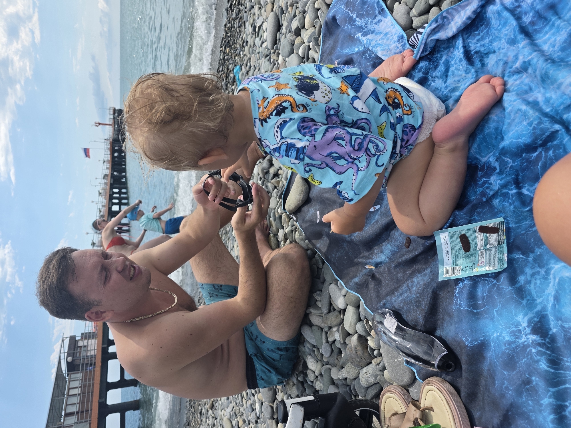 A group of people are on a pebble beach near the sea. In the foreground, a man in blue patterned swim trunks and a gold chain is sitting on a blue, wave-patterned towel, looking at a toddler. The toddler, wearing a light blue shirt with sea creatures and a diaper, is also on the towel, reaching towards something the man is holding. Nearby on the towel are a pack of candies and a pair of goggles. In the background, a pier extends into the water, with a Russian flag flying on it. Other people, including a woman in a red bikini, are visible on the beach and near the pier. The sky is partly cloudy, suggesting it is daytime with pleasant weather. The overall scene captures a casual family outing at the beach.