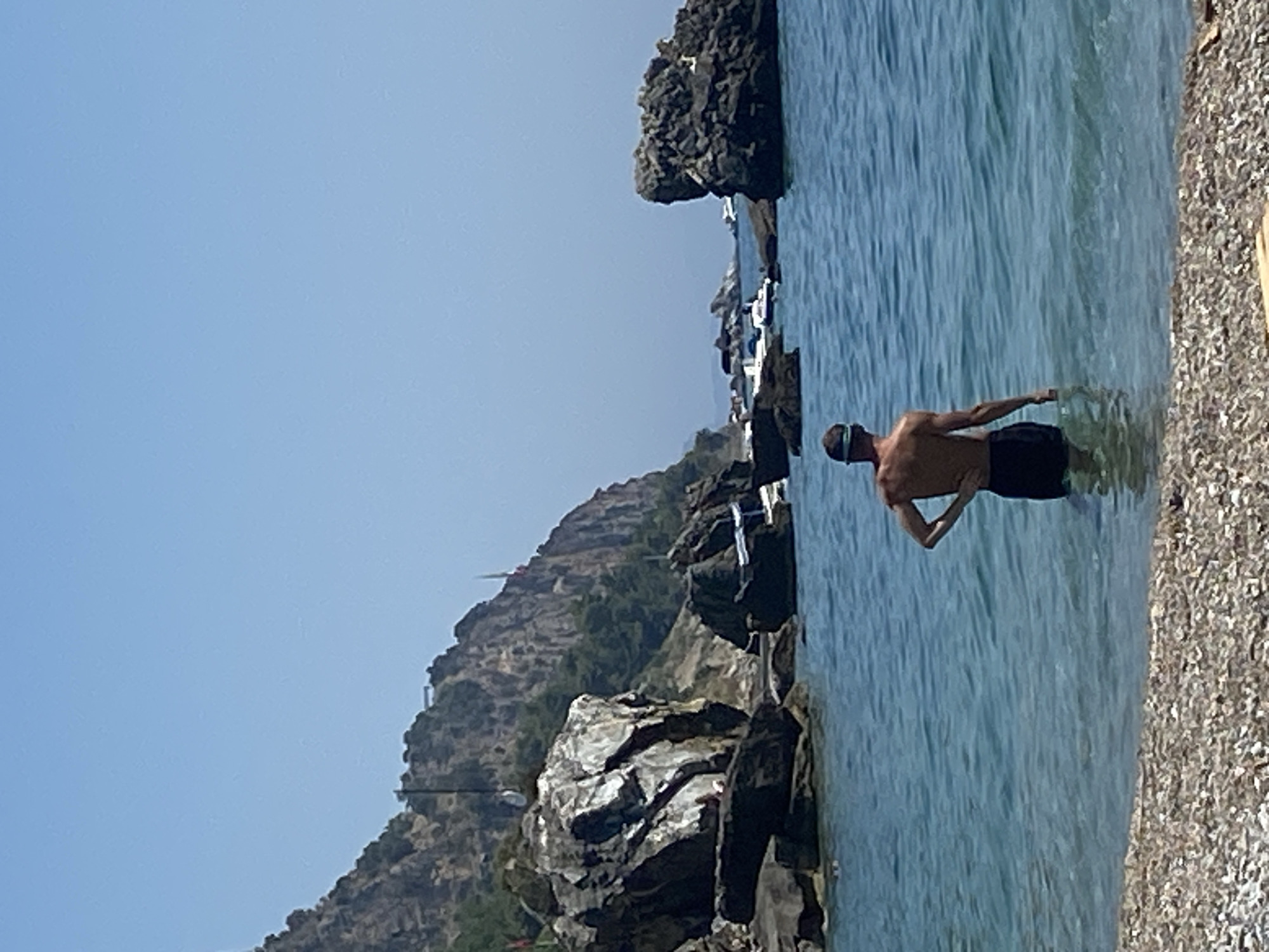 A person stands in the sea, facing away from the camera. The person is wearing dark shorts and a cap. The water is clear and blue. In the background, there are rocky cliffs covered in green vegetation. Several boats are visible on the water. The shore in the foreground is covered with pebbles. The sky is clear and blue. This scene is located at a beach in Prijevor, Montenegro.