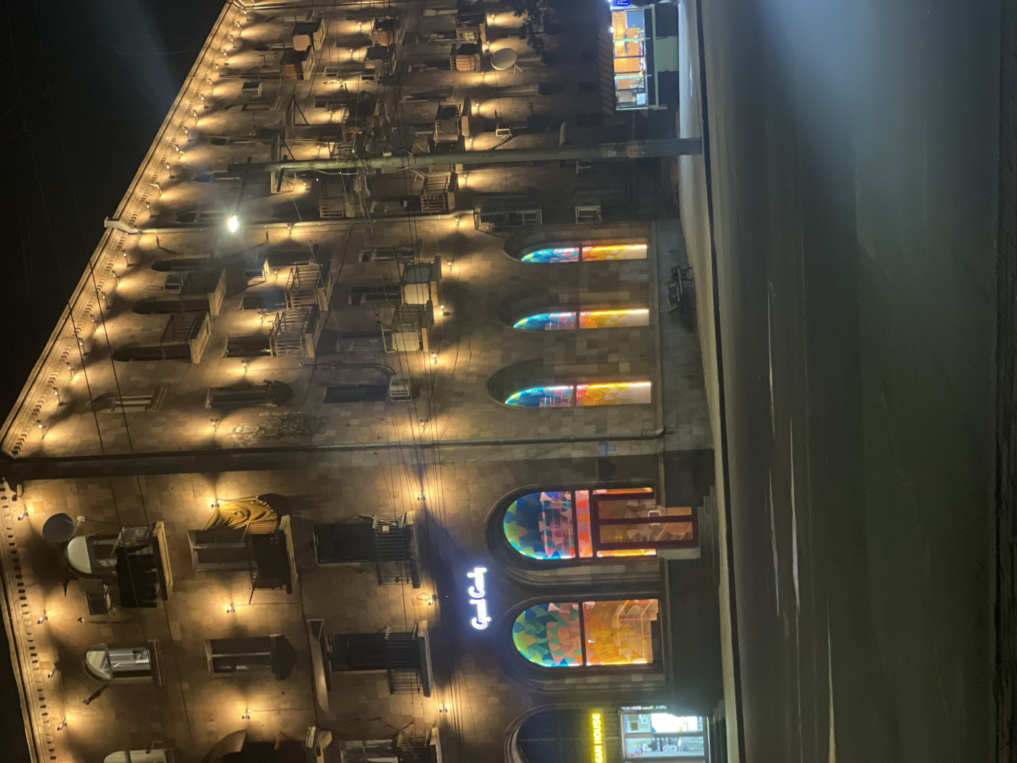The image depicts a tall building illuminated at night. The building appears to be made of stone and has multiple stories, each featuring windows and balconies. The windows are decorated with colorful, stained-glass-like patterns. The building's facade is adorned with small, evenly spaced lights that cast a warm glow. The signage "Grand Candy" is visible on the building's front. A roadway runs along the right side of the building. The location is Yerevan, Armenia.