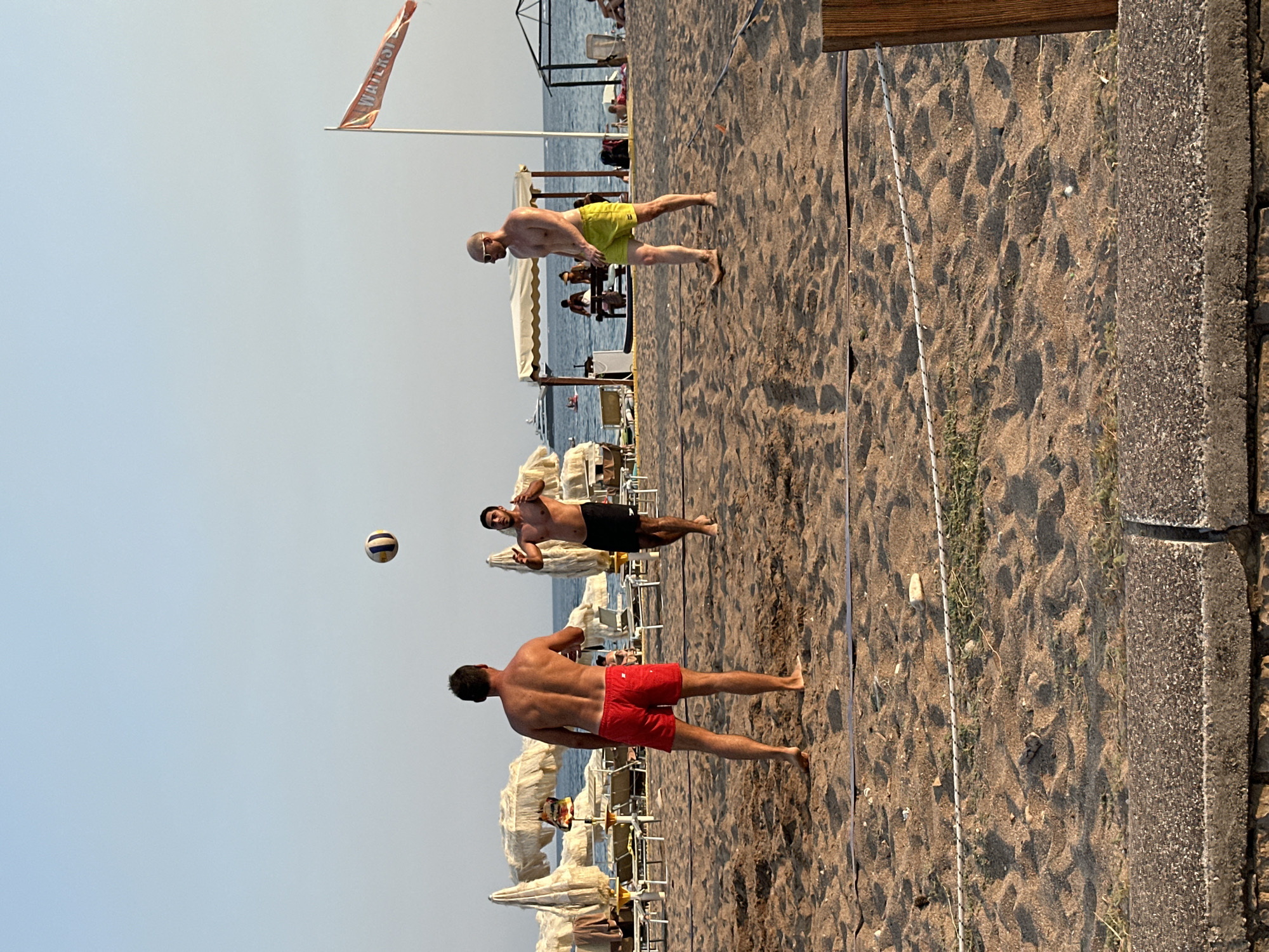 In Boreti, Montenegro, three men are playing beach volleyball on a sandy court. The man in the foreground on the left is wearing red swim trunks and facing away from the camera, his back to the viewer. In the middle, a man wearing black swim trunks is looking up at the volleyball, which is in the air above him. To the right, a bald man with sunglasses and yellow swim trunks is walking on the sand, facing towards the center of the court. In the background, beach umbrellas and lounge chairs are set up on the sand, and the sea is visible beyond. A flag with the word "WATER" is also visible in the background. The overall scene depicts a recreational activity on a sunny day at a beach resort.