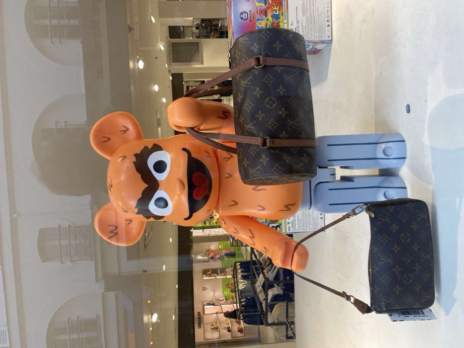 A large, orange Bearbrick figure, characterized by prominent eyebrows, large white eyes, and a wide red mouth, stands as the central subject within a retail window display in Paris, France. The figure, textured with small bumps and supported by light blue block-like legs, holds two brown Louis Vuitton monogram canvas handbags. In its raised right hand (viewer's left), it grasps the handle of a cylindrical Louis Vuitton Papillon bag. From its lower left hand (viewer's right), a thinner strap descends to a smaller, rectangular Louis Vuitton Pochette Accessoires bag resting on the white display surface.
Behind the glass, the interior of a brightly lit retail store is visible, featuring a "LONGCHAMP" brand sign on the left, alongside shelves displaying handbags and racks of clothing. Reflected in the upper portion of the display glass are architectural elements of a classic Parisian building, showing cream-colored stone facades, arched windows, and dark, ornate railings, consistent with Haussmannian design. A faint, dark reflection of a person is also present in the upper right portion of the reflection.