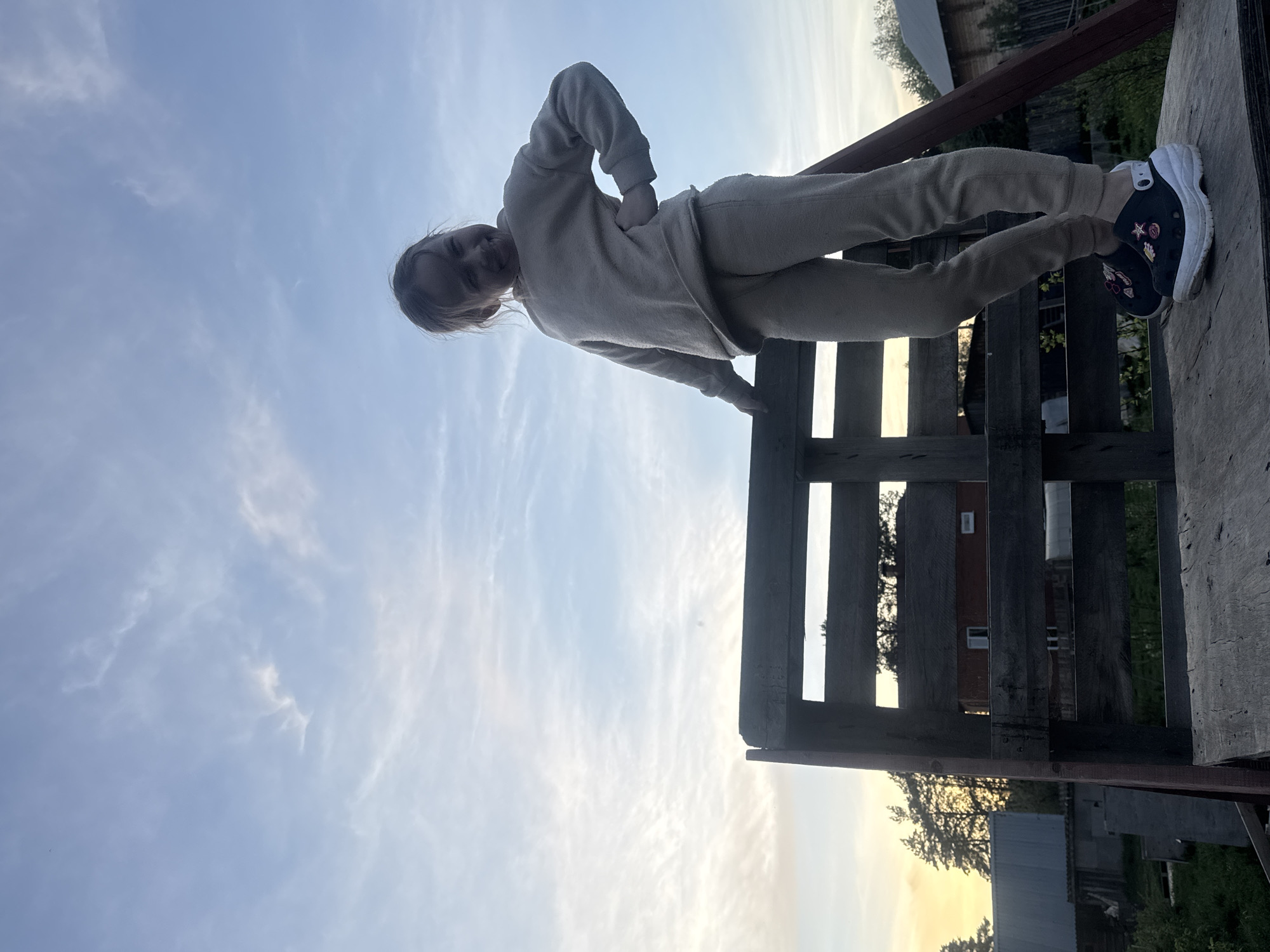 A person wearing a beige sweatsuit and black Crocs with embellishments stands on a weathered wooden structure. The structure has vertical slats and a wooden platform. The sky is visible in the background, transitioning from a light blue at the top to a yellowish-white near the horizon. Through the gaps in the wooden structure, glimpses of buildings and trees can be seen. The location is consistent with an outdoor setting in Rusalevka, Russia.