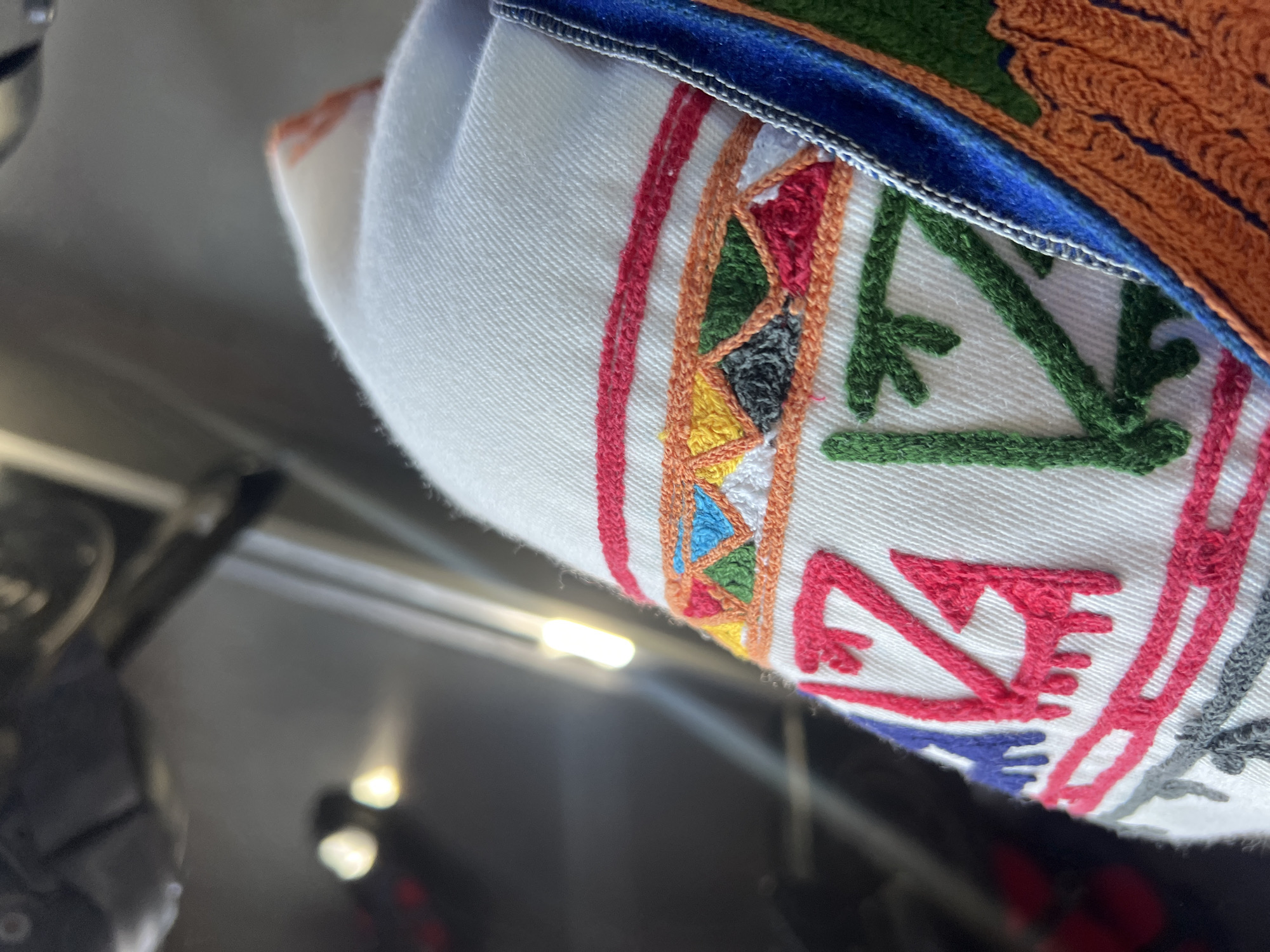 The image presents a detailed, close-up forensic view of a white textile object featuring intricate multi-colored embroidery, captured in the city of Slivovo, Bulgaria. The primary subject is a section of white woven fabric, possibly cotton or linen, characterized by a visible weave. A horizontal band of geometric embroidery adorns the upper portion, composed of small, dense stitches forming triangular and angular patterns in red, orange, yellow, dark green, light blue, dark gray, and white threads. Below this, larger, stylized patterns are embroidered in thick, raised threads: prominent red motifs resembling angular "Z" or lightning bolt shapes and triangles, flanked by similar abstract designs in dark blue to the left and forest green to the right. A distinct vertical trim of blue fabric, possibly velvet or felt, with a narrow white piping, runs along the right edge of the white textile, adjacent to a section of dense, textured orange embroidery. The background is severely out of focus, appearing as a dark, indistinct expanse dotted with several bright, blurred light sources and elongated reflections, indicative of an indoor setting with artificial illumination. No human figures or discernible actions are present in the frame.