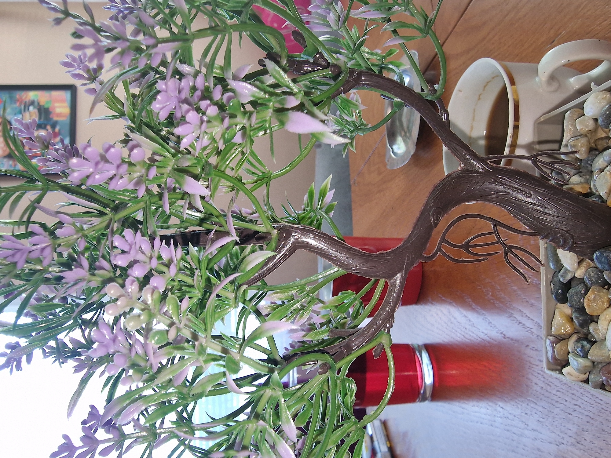 The image depicts an artificial plant with purple flowers and green foliage, situated on a wooden surface. A white ceramic mug filled with a dark liquid is visible to the right of the plant. Below the plant are two red cylindrical objects. The plant is positioned in a bed of small rocks. A silver spoon is partially visible near the mug. A framed picture can be seen on the wall in the background.