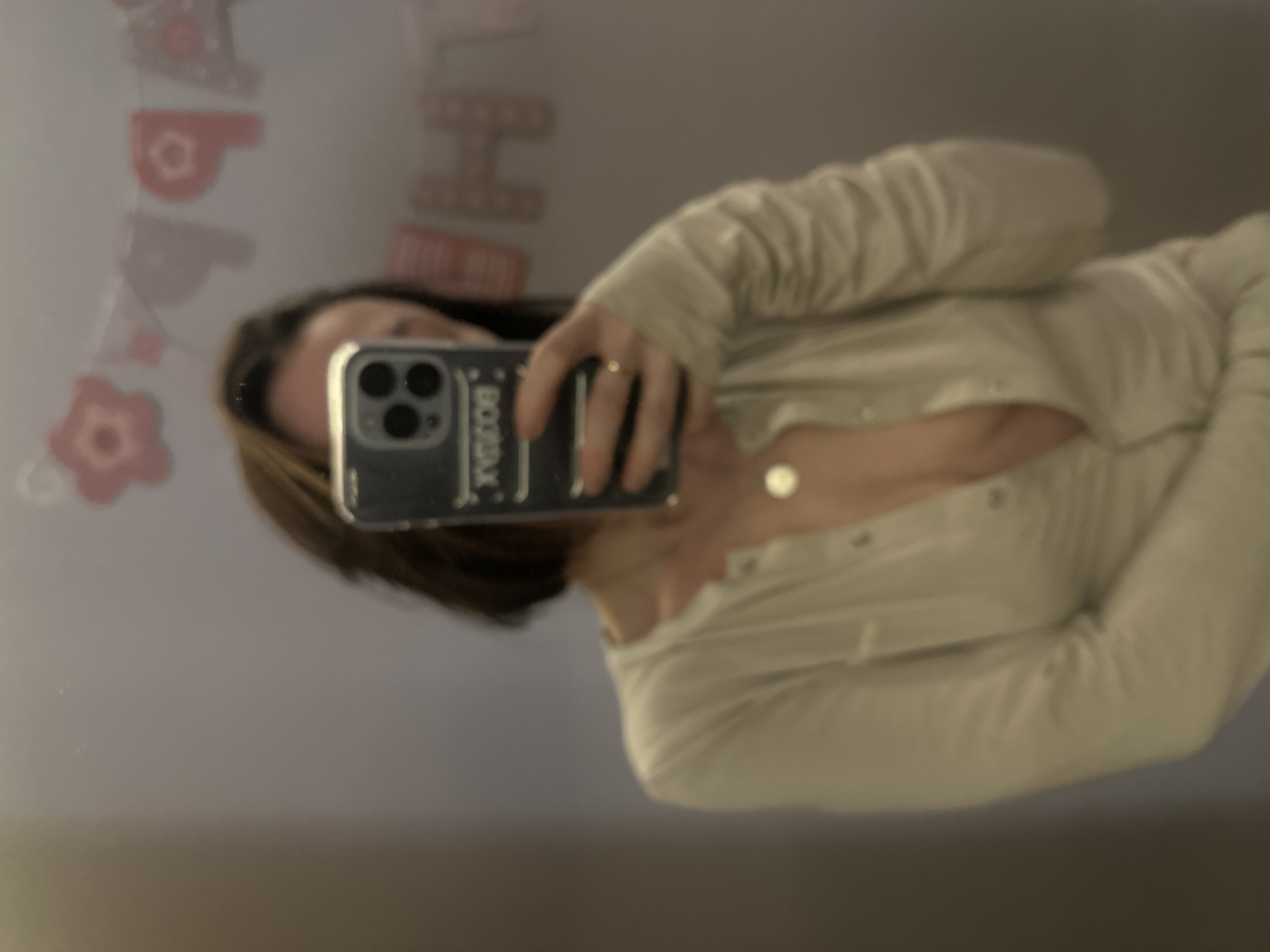 A woman is taking a selfie in front of a mirror. The mirror reflects a "Happy Birthday" banner. The woman is wearing a light-colored top with buttons and has a necklace. She is holding a cell phone to take the picture. The background is a wall, but the details are not clear. This image was not captured in Votkinsk, Russia.