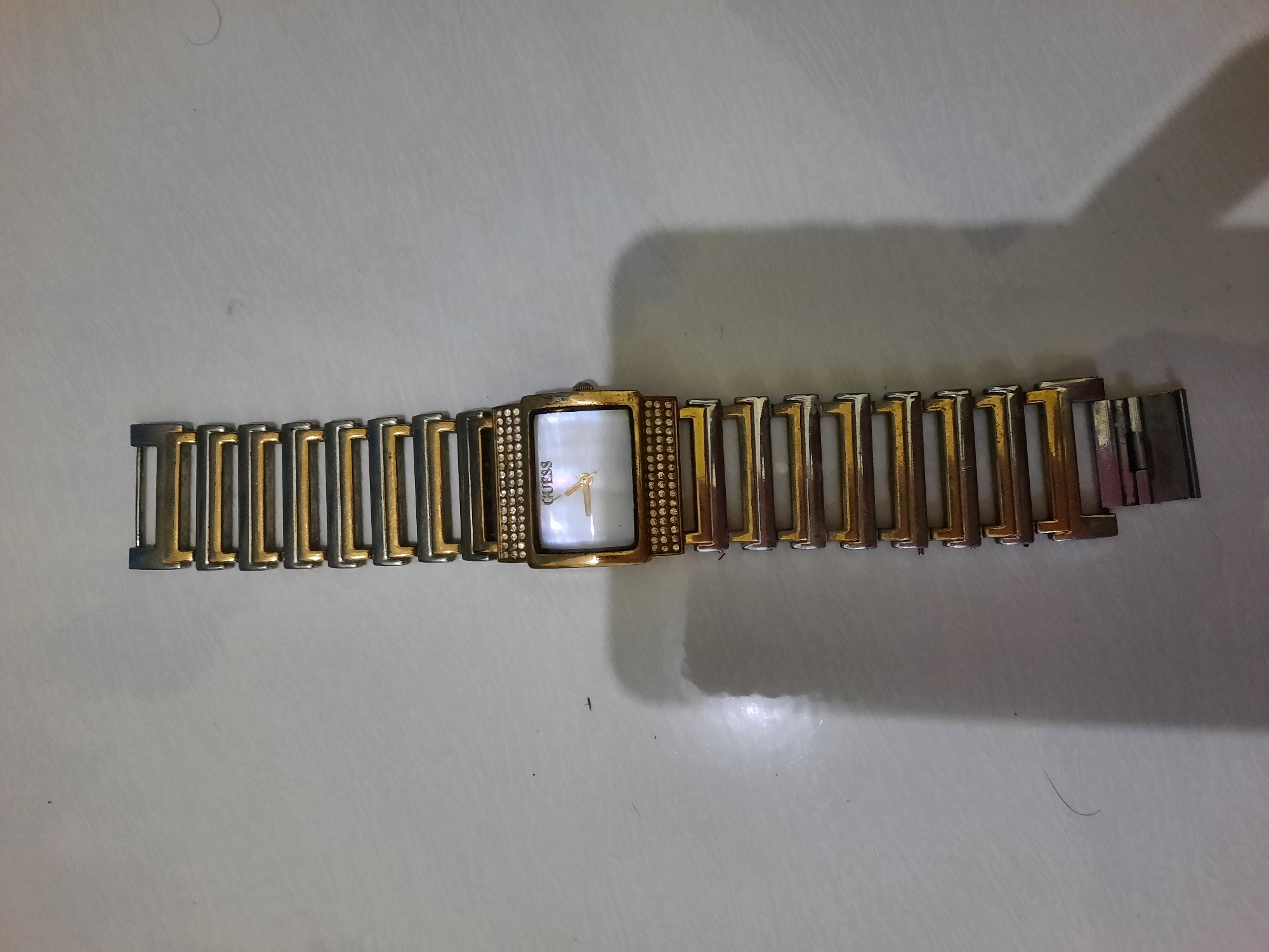 An overhead, full shot shows a women's gold and silver-toned Guess watch lying on a textured, light-colored surface. The watch is laid out flat, with the band extending upwards from the watch face. The watch face itself is rectangular with rounded corners and has a mother-of-pearl dial. Gold-colored hour and minute hands are visible, and the word "GUESS" is written in black above the hands. The bezel of the watch is encrusted with small, clear rhinestones. The band is made of alternating gold and silver-toned rectangular links. The watch is positioned vertically in the center of the frame, with its clasp at the bottom. A soft shadow falls to the left of the watch. The image was taken in Lhokseumawe, Indonesia.