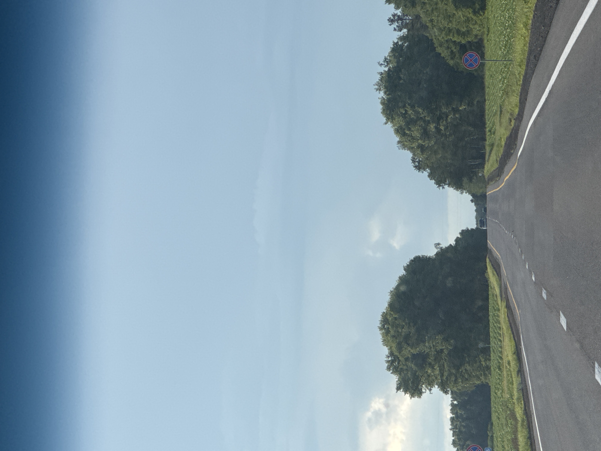 This is a landscape photograph taken from a moving vehicle on a paved road, possibly a highway, in Belarus. The road is marked with a white line separating lanes and a yellow line along the shoulder. The right side of the image shows the road curving to the right, bordered by a grassy shoulder and a dense line of lush green trees. In the distance, more trees and a few vehicles can be seen on the road. There is a traffic sign on a pole visible on the left side of the road, indicating a no parking zone. The sky is a pale blue with scattered white clouds, suggesting it is daytime, likely late morning or afternoon. The image has a bright and airy feel, with the sun casting a slight glare on the road. The left side of the image transitions into a gradient of blue, suggesting the sky or possibly a reflection. The overall scene is serene and rural, with the focus on the natural environment and the road infrastructure.