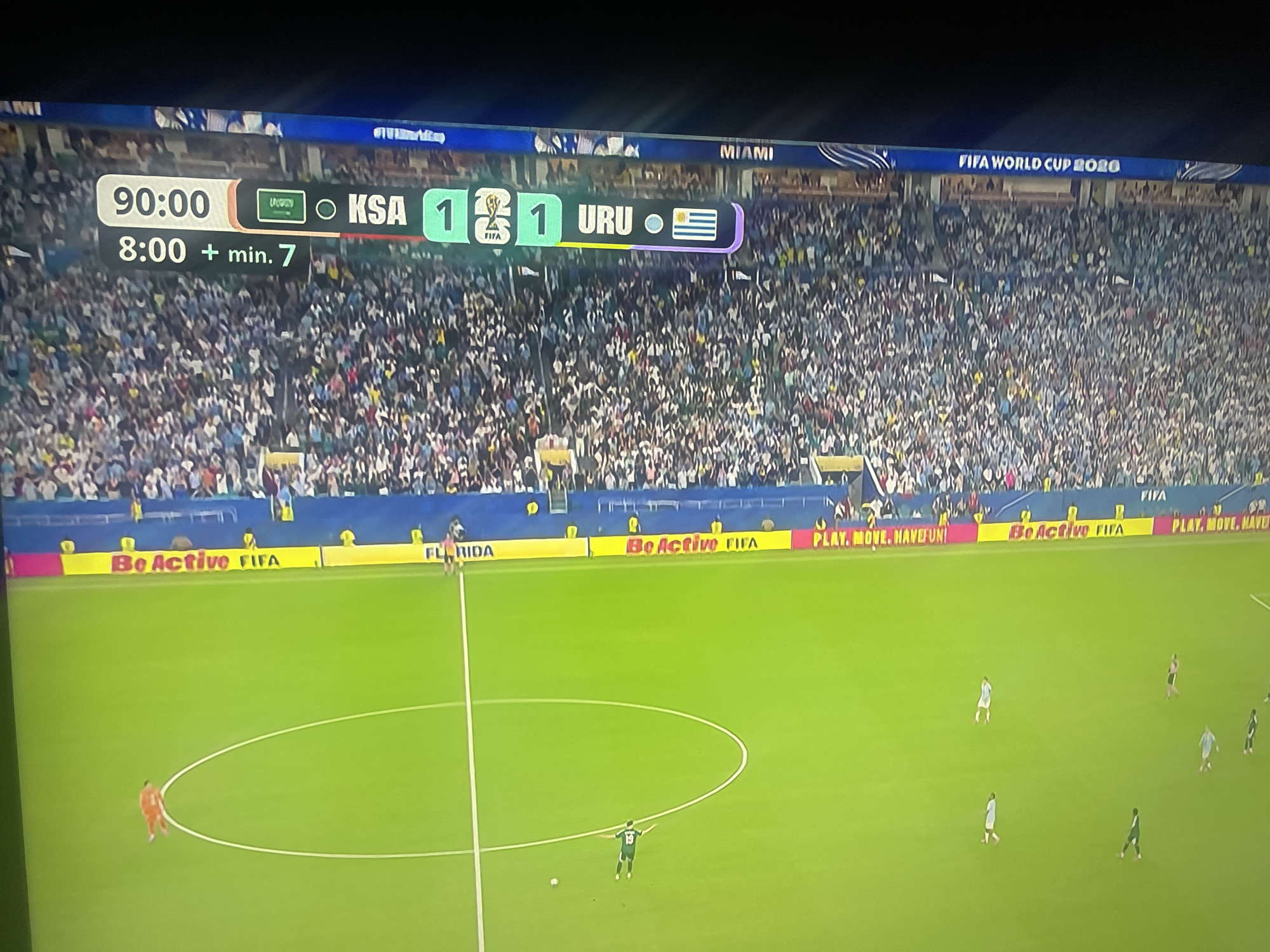The image shows a soccer match scoreboard displaying "90:00" indicating the end of the 90 minutes of play, and "8:00 + min. 7" suggesting 7 minutes of added time. The score is KSA 1 - 1 URU. The stadium is filled with spectators. The field is green, with white lines marking the boundaries and a center circle. There are several players on the field, including a goalkeeper in orange and other players in white and green uniforms. Advertising banners are visible around the perimeter of the field, including "Be Active FIFA" and "PLAY. MOVE. HAVE FUN." The header of the scoreboard displays "MIAMI" and "FIFA WORLD CUP 2020". The scene is a stadium during a soccer match. There is no information to suggest the city is Ruse, Bulgaria.