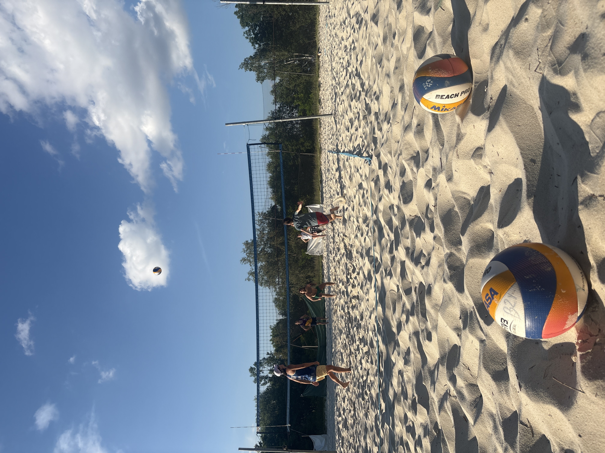 A group of people are playing volleyball on a sandy court. The sky is blue with scattered white clouds. In the foreground, two volleyballs are resting on the sand. In the middle ground, several individuals are actively engaged in the game, with one person jumping to hit the ball over the net. The net is stretched across the court, supported by poles. Trees line the background of the scene. The lighting suggests it is daytime, likely late afternoon due to the shadows.
