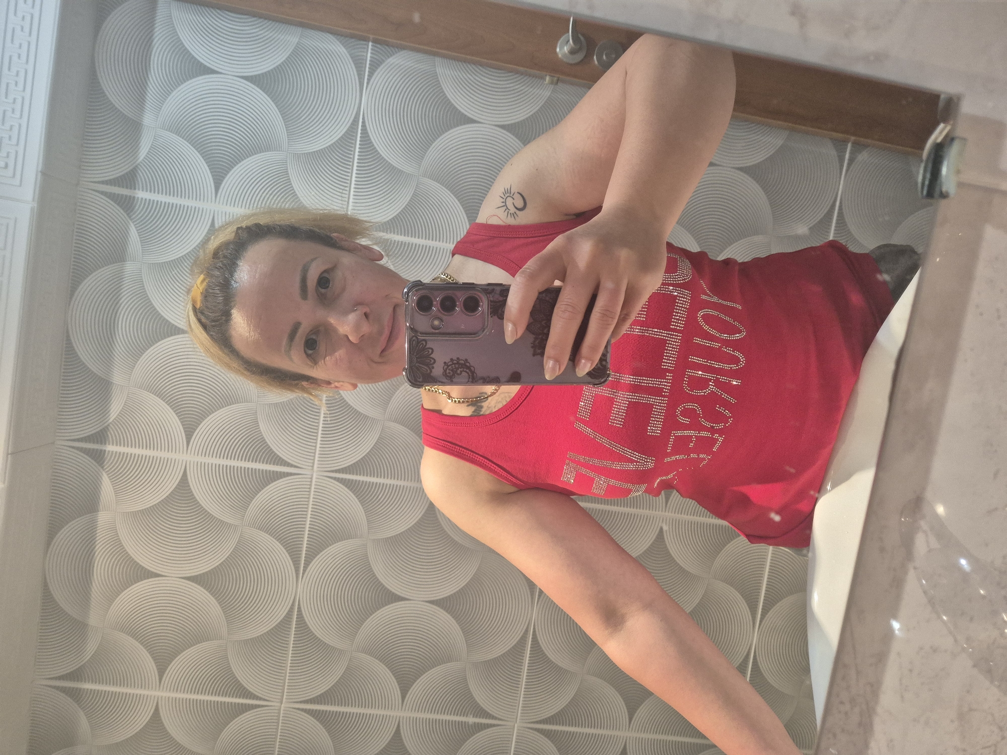 A woman is depicted taking a mirror selfie in what appears to be a bathroom. She has short, blonde hair, a small stud piercing on her nose, and a gold chain necklace. On her upper left arm, there is a tattoo of a sun and a crescent moon. She is wearing a red sleeveless top adorned with sparkling text that, when mirrored, reads "VICTORIA'S SECRET."
She holds a dark-colored smartphone, visible with its three camera lenses, encased in a decorative, patterned cover, using it to capture her reflection. The background shows a distinctive wall covered in grey and white tiles featuring a repeating pattern of concentric semicircles. A portion of a white sink is visible at the bottom right of the frame, and a wooden border frames the mirror at the top. A silver faucet handle is also reflected in the mirror. The image is oriented sideways, suggesting it was captured with the phone held horizontally.