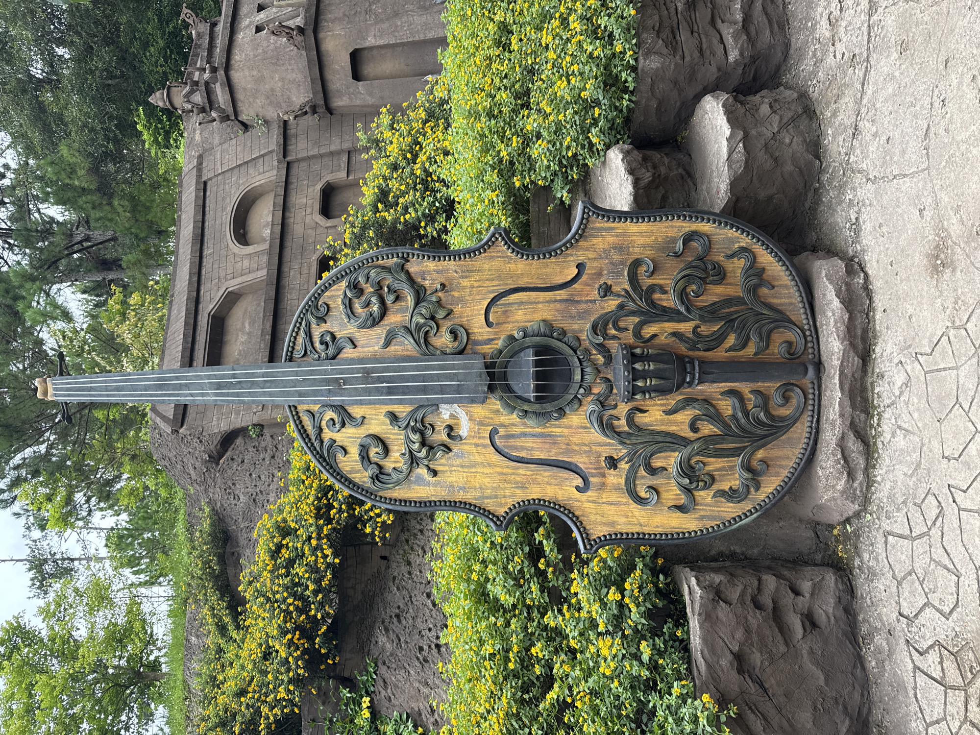 This image captures a large, ornate cello sculpture positioned outdoors. The cello is made of wood with dark, decorative scrollwork. It stands in front of a textured stone wall and lush green foliage with small yellow flowers. Behind the foliage, a section of a stone building with arched windows and decorative architectural elements is visible. The ground in the foreground is paved with irregular stone tiles. The overall setting suggests a garden or park environment. The scene is located at the Da Lat Flower Park in Da Lat, Vietnam, a city known for its vibrant floral displays and European-inspired architecture. There are no people or animals present in the image. The lighting suggests it is daytime with an overcast sky.