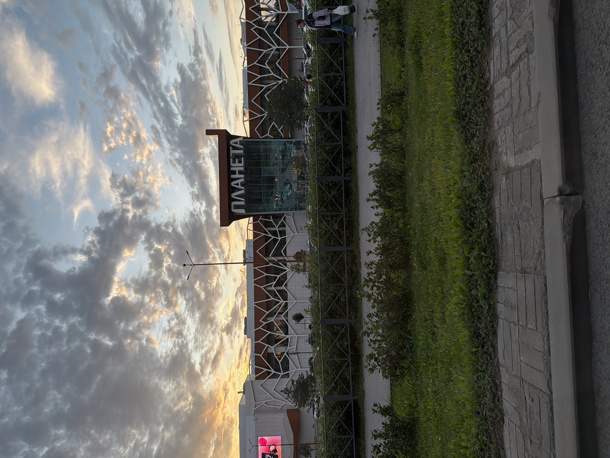 The image captures the exterior of the "Planeta" shopping mall in Perm, Russia, during what appears to be sunset. The sky is filled with dramatic, cloudy formations, with patches of orange and yellow light indicating the setting sun, particularly on the left side of the frame. The shopping mall's modern architecture is visible, featuring a large glass facade and a distinctive white geometric pattern overlaying the building's structure. The name "ПЛАНЕТА" is prominently displayed in large letters on the building.
In the foreground, a paved sidewalk runs horizontally across the bottom of the image. Beyond the sidewalk, a strip of green grass and dense bushes is visible, separated from the mall by a fence. Cars are parked in a lot behind the fence and in front of the mall.
A single person is walking along the sidewalk from right to left. They are wearing a jacket and jeans, and carrying a white plastic bag in their right hand. The person's gait suggests they are casually strolling. The overall atmosphere is calm and serene, characteristic of the evening hours in a suburban or city environment.